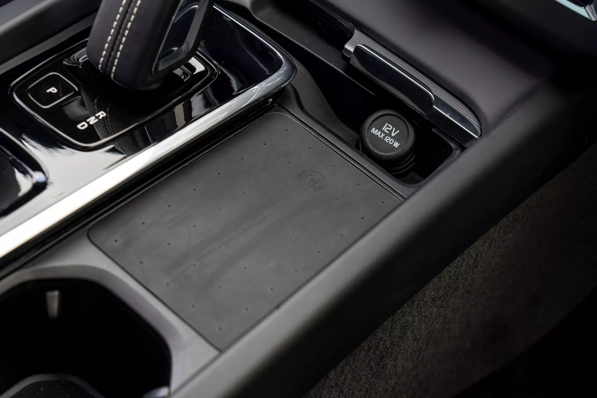 Zbliżenie pod kątem na obszar konsoli środkowej w Volvo XC60 2021 (druga generacja, lifting) z perspektywy pasażera, z automatyczną dźwignią zmiany biegów w pozycji 'park', antypoślizgowym schowkiem, przełącznikiem elektronicznego hamulca postojowego, uchwytami na kubki i kombinacją miękkich w dotyku plastikowych i metalowych wykończeń w przytłumionych odcieniach ciemnej szarości i czerni.