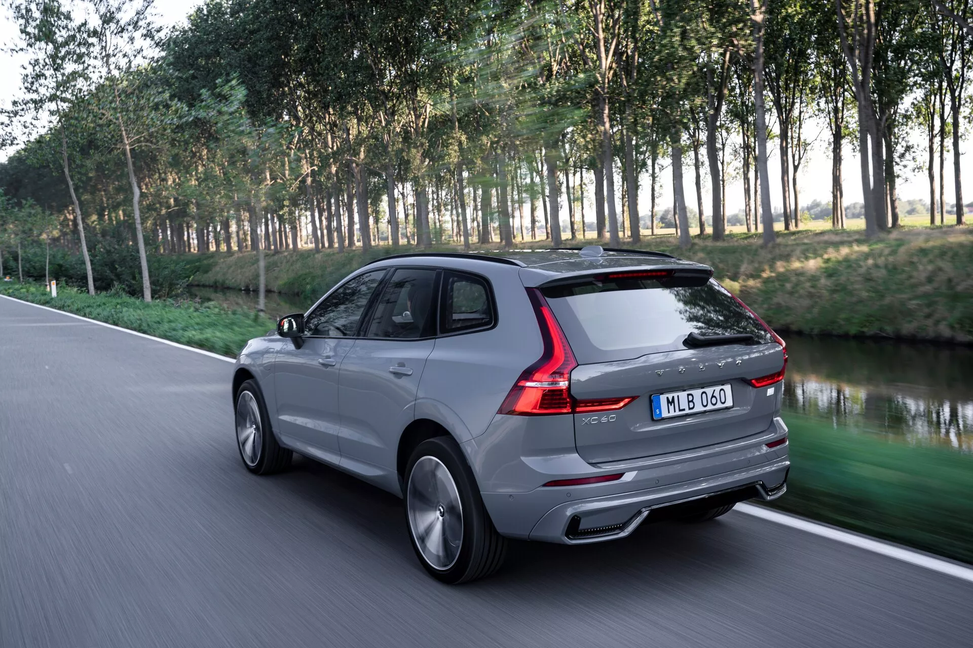 Dynamiczny widok z tyłu na trzy czwarte nowoczesnego SUV-a Volvo XC60 2021 (2. generacja - lifting) na asfaltowej drodze, podkreślający elegancki design tylnej części, w tym światła tylne, tylną szybę, bagażnik i linię dachu. Obraz, wykonany z lekko obniżonej perspektywy, pokazuje stylowe koła i boczne linie charakterystyczne. SUV jest pokazany w ruchu z rozmytym naturalnym tłem drzew i trawy, wskazującym na ustawienie parkowe. Srebrnoszare luksusowe pojazdy posiadają podwójne rury wydechowe, aerodynamiczny design i błyszczące wykończenie, z aluminiowymi kołami i prawdopodobnie zaawansowaną technologią, taką jak tylne światła LED.