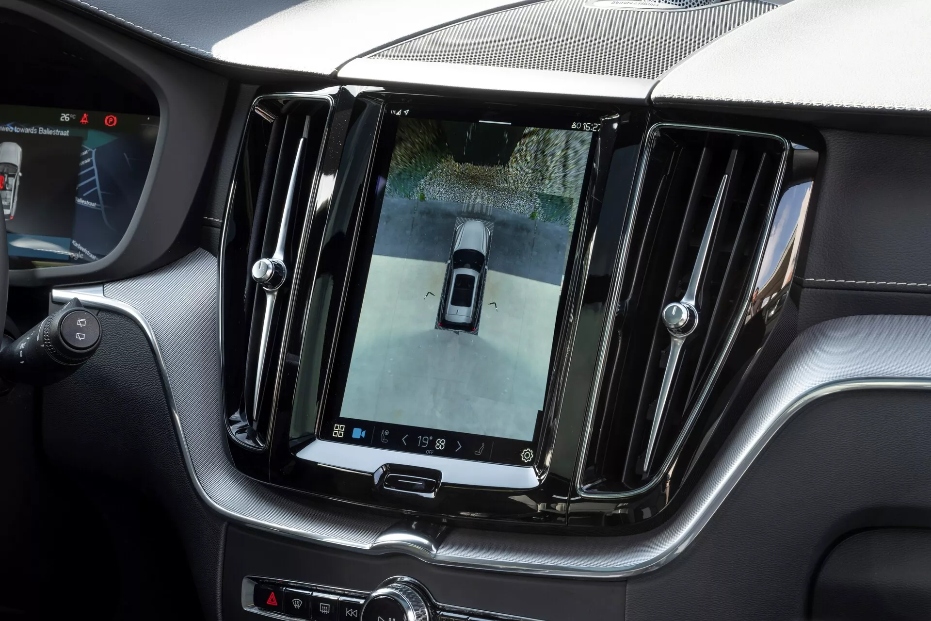 Wnętrze Volvo XC60 2021 (lifting 2. generacji) z dużym, pionowo zorientowanym ekranem dotykowym infotainment na konsoli centralnej, wyświetlającym widok z kamery cofania z perspektywy ptaka. Luksusowe wykończenie z chromowanymi akcentami i eleganckimi elementami designu, dodatkowe kontrolki poniżej ekranu i częściowy widok na kierownicę, wskazujące na wnętrze premium i zaawansowaną technologię.