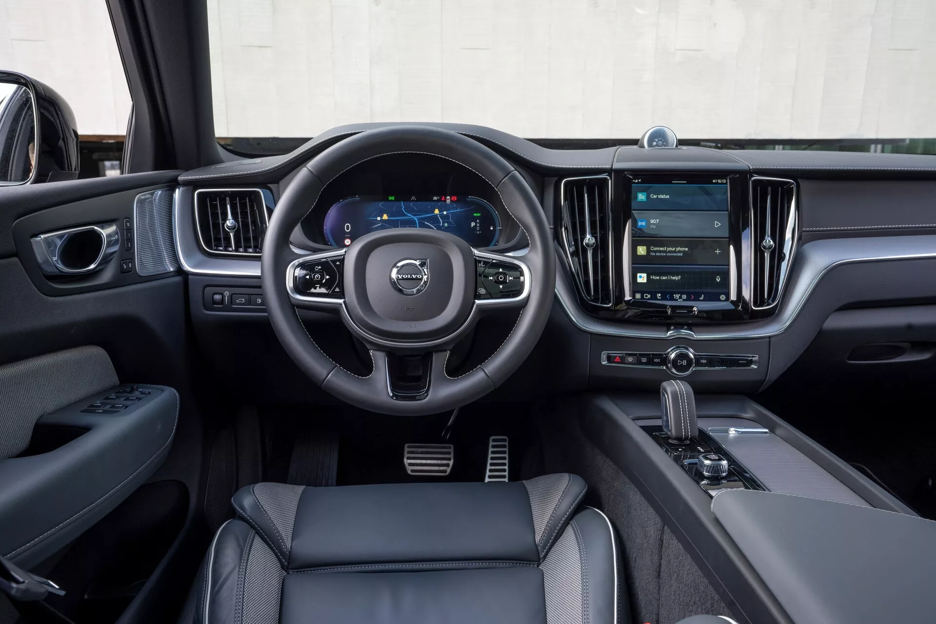 Wnętrze Volvo XC60 2021 (facelifting 2. generacji) z perspektywy kierowcy. Obraz podkreśla trójramienną kierownicę z przyciskami do sterowania dźwiękiem, komunikacją i ustawieniami tempomatu, cyfrowy zestaw wskaźników wyświetlający prędkościomierz i nawigację, oraz duży pionowy system informacyjno-rozrywkowy z ekranem dotykowym. Nowoczesne elementy wykończenia obejmują skórzane lub skóropodobne materiały, chromowane akcenty oraz automatyczny drążek zmiany biegów ze wskaźnikiem trybu jazdy. Na drzwiach kierowcy znajdują się widoczne panele sterowania szybami i lusterkami, co podkreśla technologiczne i luksusowe cechy.