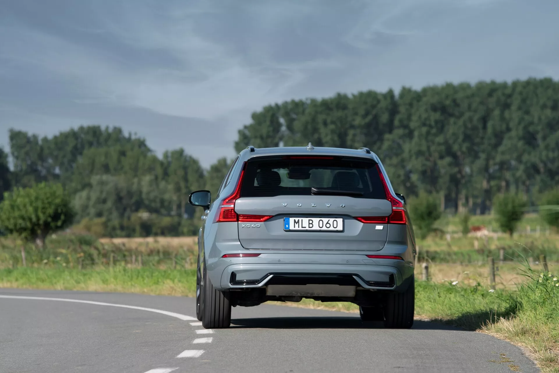 Tylny widok nowoczesnego SUV-a Volvo XC60 2021 (2. generacja-facelifting) na dwupasmowej drodze w naturalnym, wiejskim otoczeniu, pokazującego charakterystyczne tylne światła, podwójny wydech, tylny spojler z zintegrowanymi światłami hamowania, kamerę cofania, tylna wycieraczka i czujniki parkowania. Umieszczony nieco na prawo od linii środkowej, z czystym niebem i drzewami w tle.