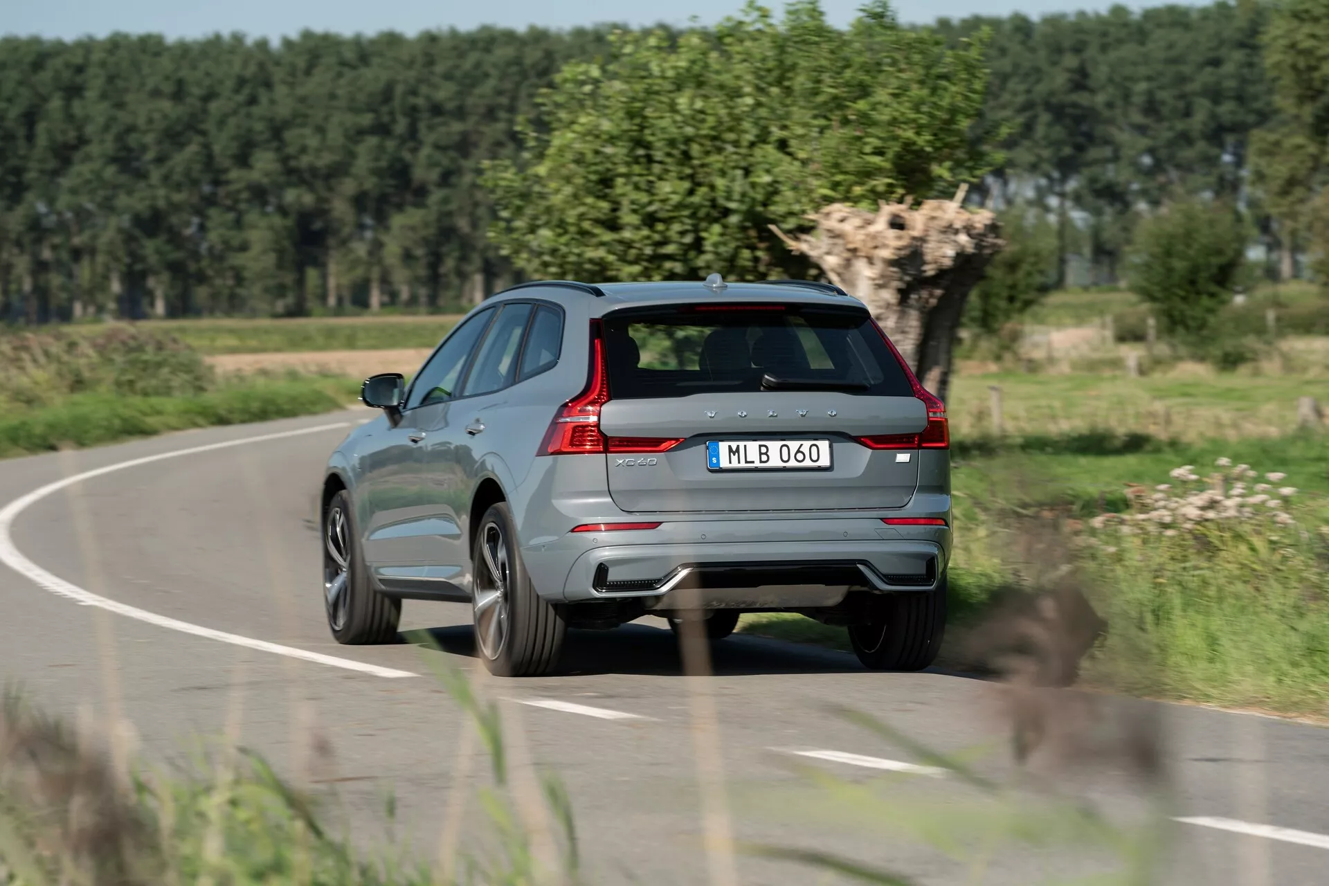 Widok tylno-trzeci ćwiartki nowoczesnego, metalicznego szarego SUV-a Volvo XC60 2021 w ruchu na wiejskiej drodze, pokazującego charakterystyczne tylne światła, podwójny układ wydechowy, tylne wycieraczki, wyraźny tylny spojler, zintegrowane czujniki w tylnym zderzaku, centralnie umieszczoną tablicę rejestracyjną, aluminiowe felgi, ostrą linię charakterystyczną wzdłuż boku, lśniące wykończenia okien i system szyn dachowych. Tło z rozmytą roślinnością i czyste niebo wskazuje na ruch i wiejski krajobraz.