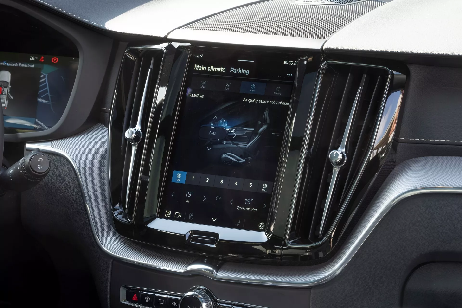 Wnętrze Volvo XC60 2021 (2. generacja z faceliftingiem) skupiające się na konsoli środkowej. Zdjęcie uchwytuje pionowo usytuowany ekran dotykowy systemu infotainment w chromowanej obudowie, wyświetlający ustawienia klimatyzacji i zakładkę wspomagania parkowania. Luksusowa mieszanka skórzanego i węglowego wykończenia otacza ekran, wraz z hybrydowym interfejsem sterowania za pomocą fizycznych przycisków i pokręteł. Zdjęcie podkreśla zaawansowany system infotainment, kontrolę klimatu, wspomaganie parkowania oraz luksusowy design wnętrza.