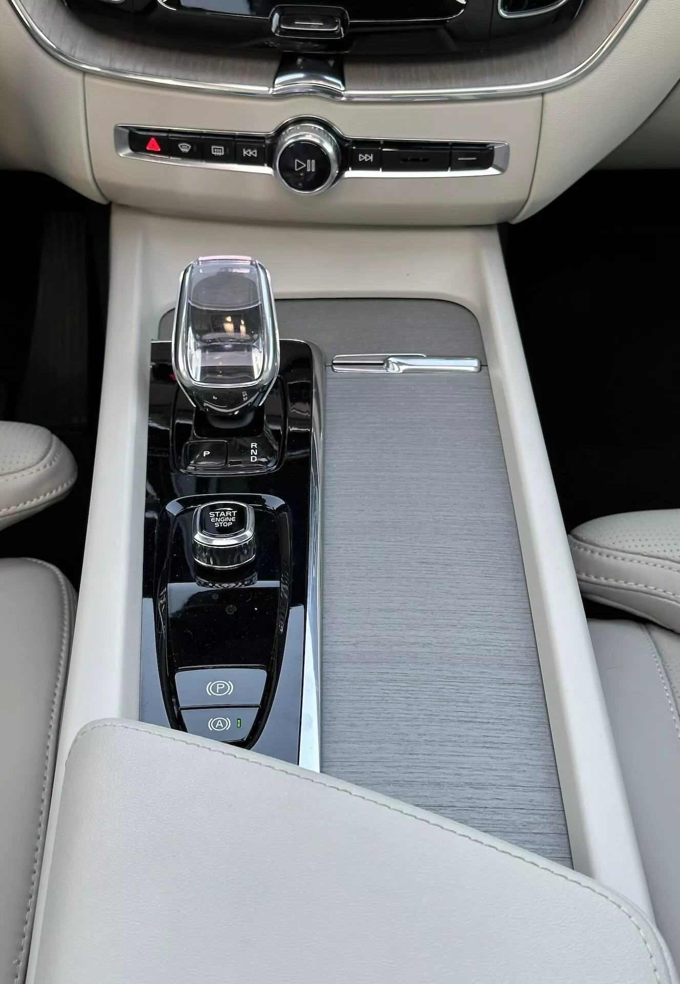 Zbliżenie na konsolę środkową w Volvo XC60 Recharge plug-in hybrydowym z 2021 roku, z beżowym skórzanym podłokietnikiem, ślizgowym wykończeniem z metalu szczotkowanego, nowoczesnym kontrolerem systemu infotainment, selektorem biegów ze szkła kryształowego dla automatycznej skrzyni biegów, oraz kontrolkami do startu/stopu silnika, wyłączania kontroli trakcji i wyboru trybu dynamiki jazdy, a także z dwustrefową kontrolą klimatu.