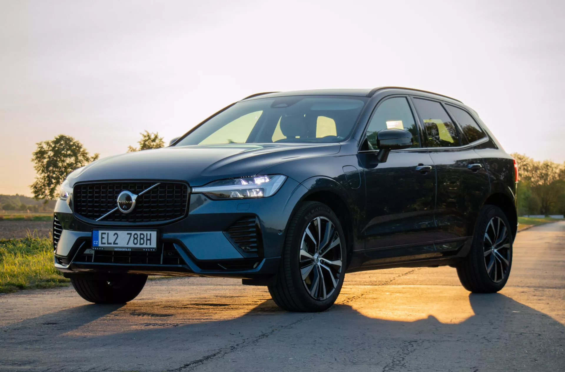 Czarne, nowoczesne SUV Volvo XC60 Recharge Plug-in Hybrid 2021 zaparkowane na asfaltowej drodze o wschodzie lub zachodzie słońca, prezentujące jego design z naciskiem na grill, reflektory LED, koła ze stopów lekkich i profil boczny na tle gradientowego nieba