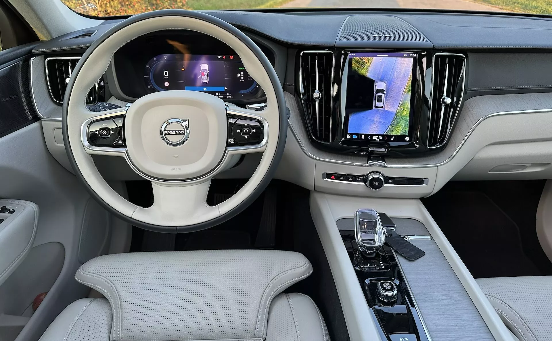 Widok wnętrza Volvo XC60 Recharge Plug-in Hybrid 2021 z tylnego siedzenia, pokazujący deskę rozdzielczą, cyfrowe wyświetlacze, kierownicę wielofunkcyjną i konsolę środkową