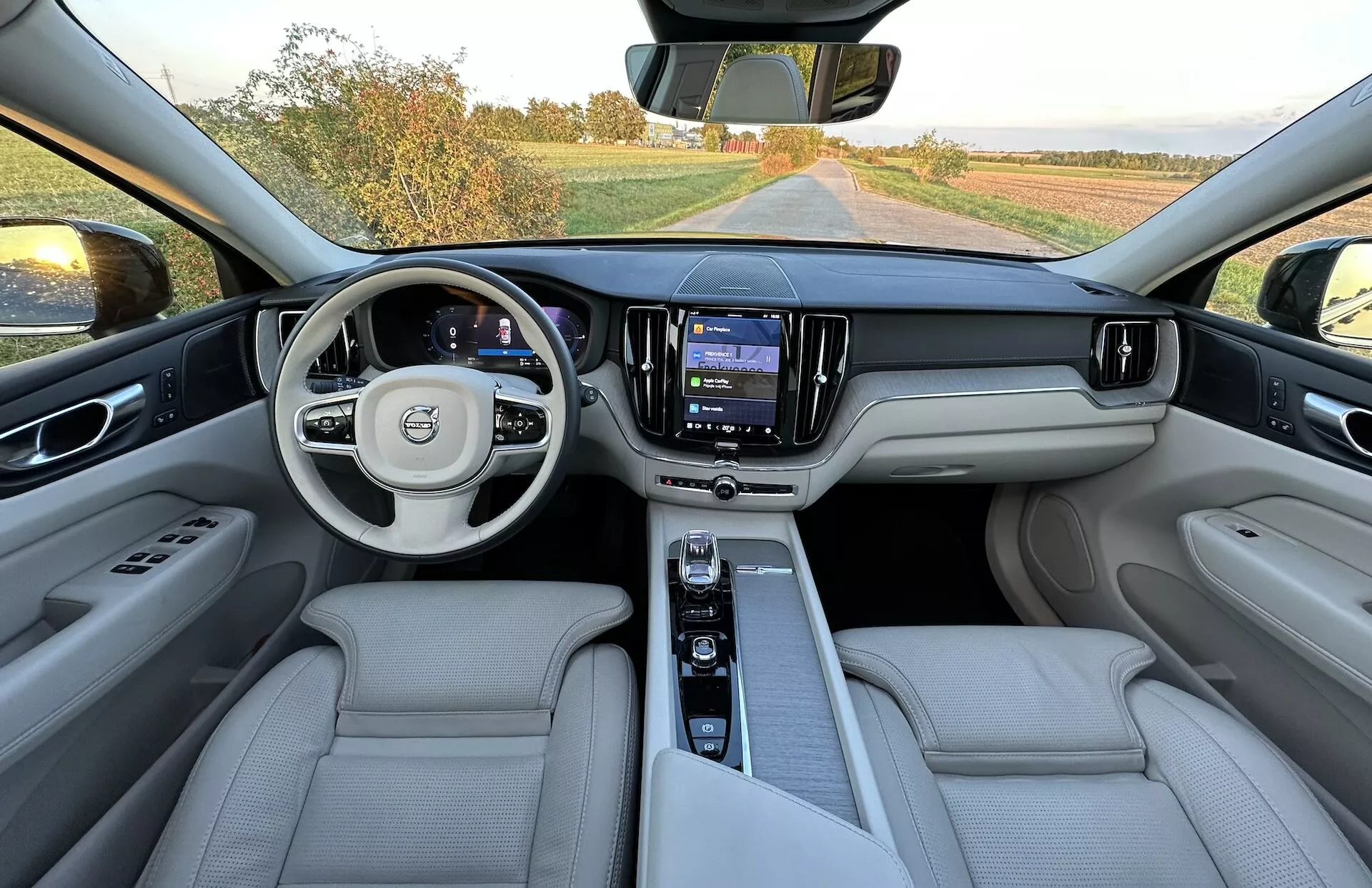 Widok wnętrza Volvo XC60 Recharge Plug-In Hybrid 2021 z tylnej części siedzeń pokazujący całą deskę rozdzielczą, wielofunkcyjną kierownicę z logo, cyfrowy panel wskaźników, centralny system informacyjno-rozrywkowy na dotykowym ekranie, i luksusowe siedzenia z jasnej skóry.