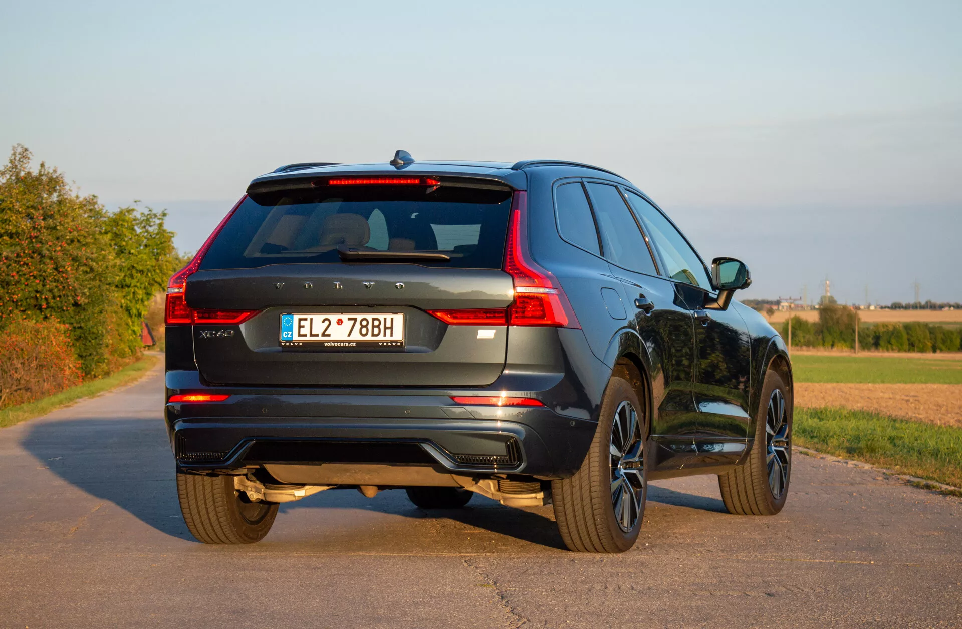Zaparkowany SUV Volvo XC60 Recharge plug-in hybrydowy z 2021 roku na utwardzonej drodze, prezentujący tylną część pojazdu z perspektywy trzech czwartych, podkreślający diodowe tylne światła, aluminiowe felgi i dynamiczny design na tle wiejskiego krajobrazu pod bezchmurnym niebem