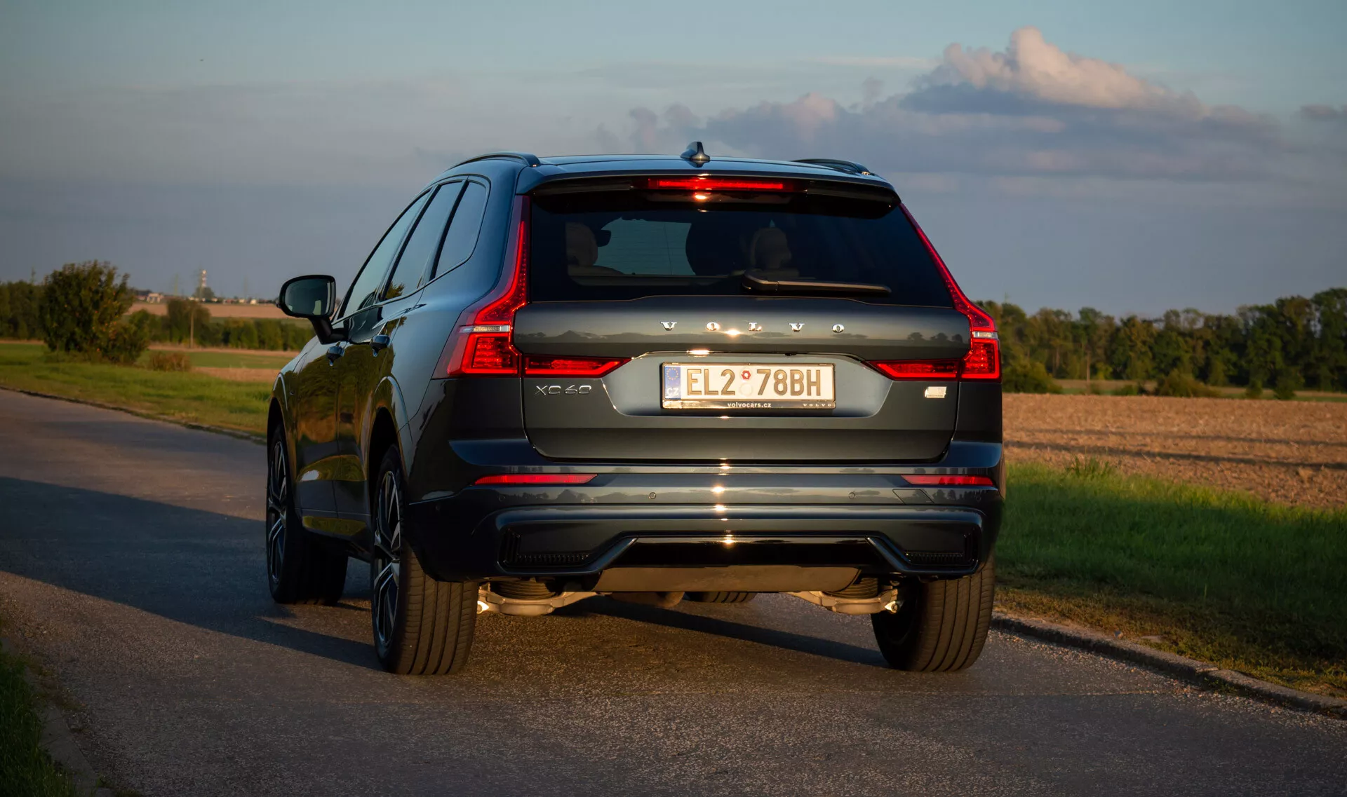 SUV Volvo XC60 Recharge plug-in hybrydowy 2021 na asfaltowej drodze, uchwycony z tylnego kąta trzech czwartych podkreślający LEDowe tylne światła pojazdu, system wydechowy i akcenty chromowane podczas późnego popołudnia