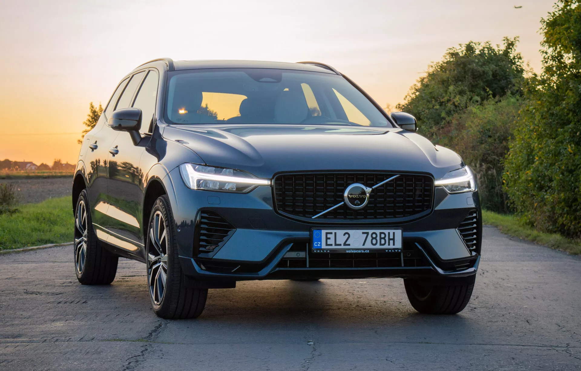 Luksusowy SUV Volvo XC60 Recharge Plug-In Hybrid 2021 na utwardzonej drodze z naturalnym krajobrazem podczas złotej godziny, prezentujący przedni grill, reflektory LED, rzeźbioną maskę, koła z lekkich stopów o wielu szprychach i błyszczące wykończenie