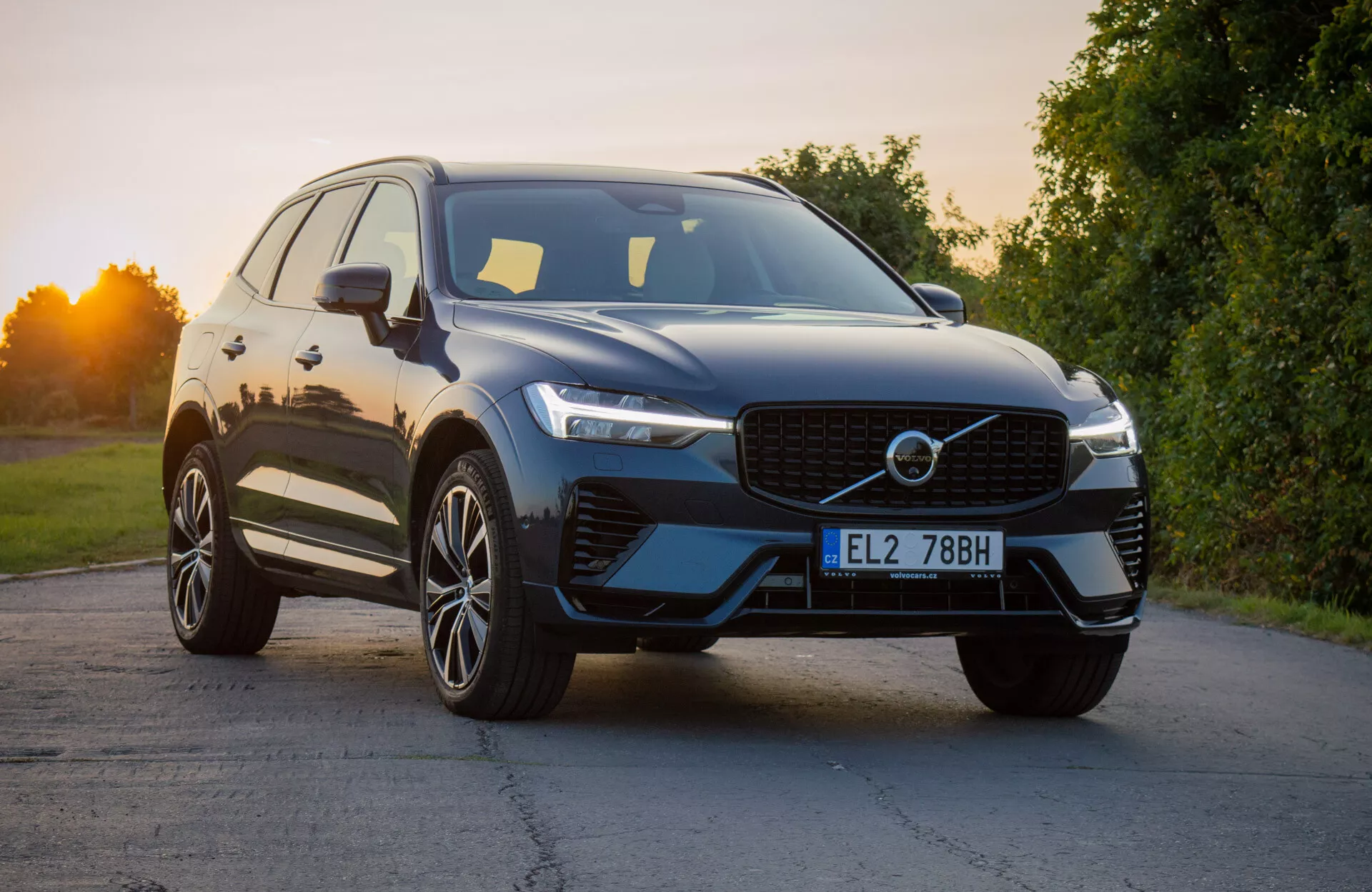 Volvo XC60 Recharge Plug-In Hybrid 2021 w nowoczesnym designie SUV, lekko pochylony aby podkreślić przednią ćwiartkę, ukazującą charakterystyczną kratę, smukłe reflektory LED i aerodynamiczny przedni zderzak z zintegrowanymi wlotami powietrza, uzupełnione o błyszczące wykończenie zewnętrzne i premium koła ze stopu z wieloma szprychami pod naturalnym światłem dziennym.