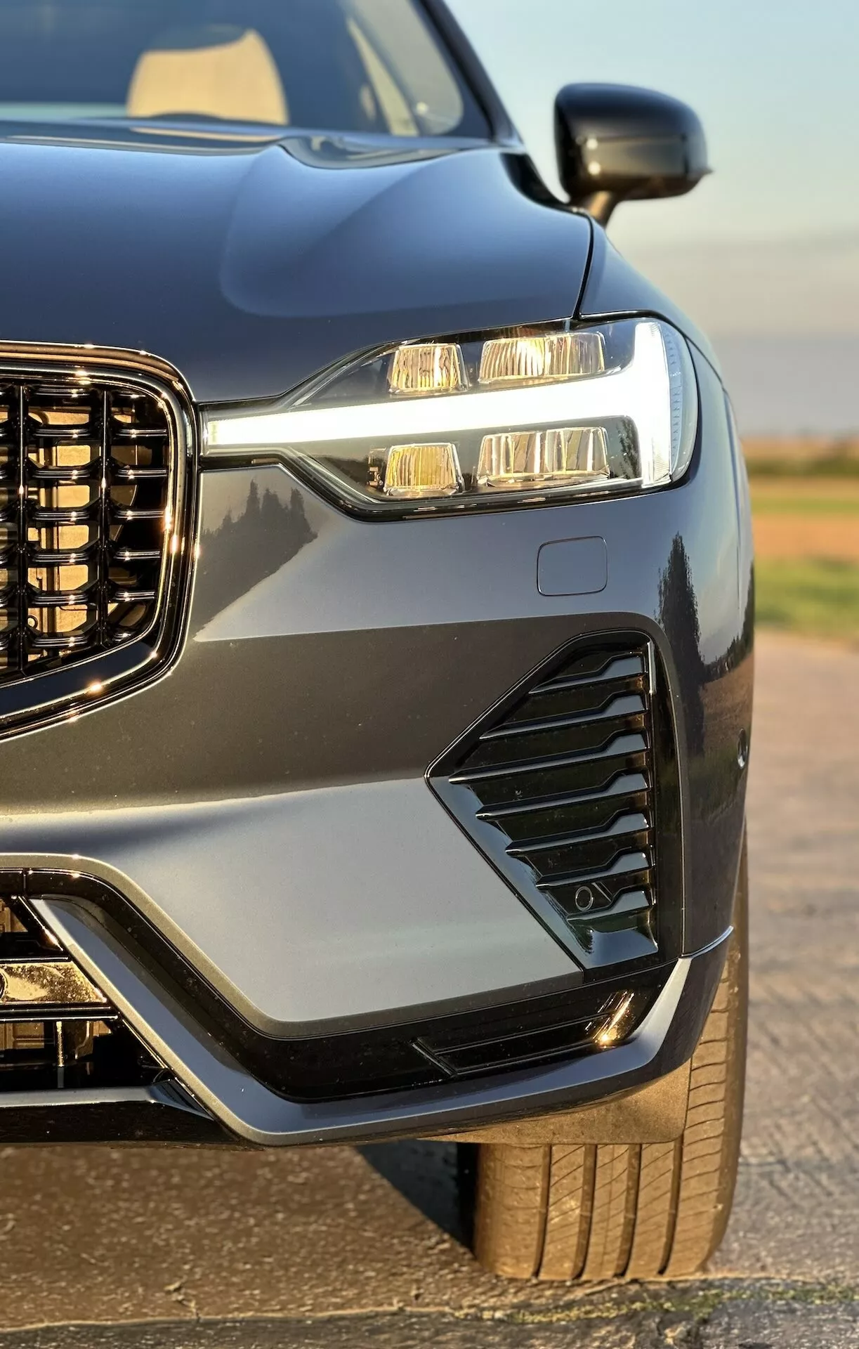 Zbliżenie przedniej części bocznej Volvo XC60 Recharge Plug-In Hybrid 2021, podkreślającej pionową kratę, LEDowe reflektory z światłami do jazdy dziennej, stylizowane wloty powietrza i światła przeciwmgielne, sportową maskę i błotniki, metaliczny lakier z kontrastującymi listwami oraz wgląd w jego solidne koło i oponę, podkreślone połyskliwym wykończeniem pojazdu.