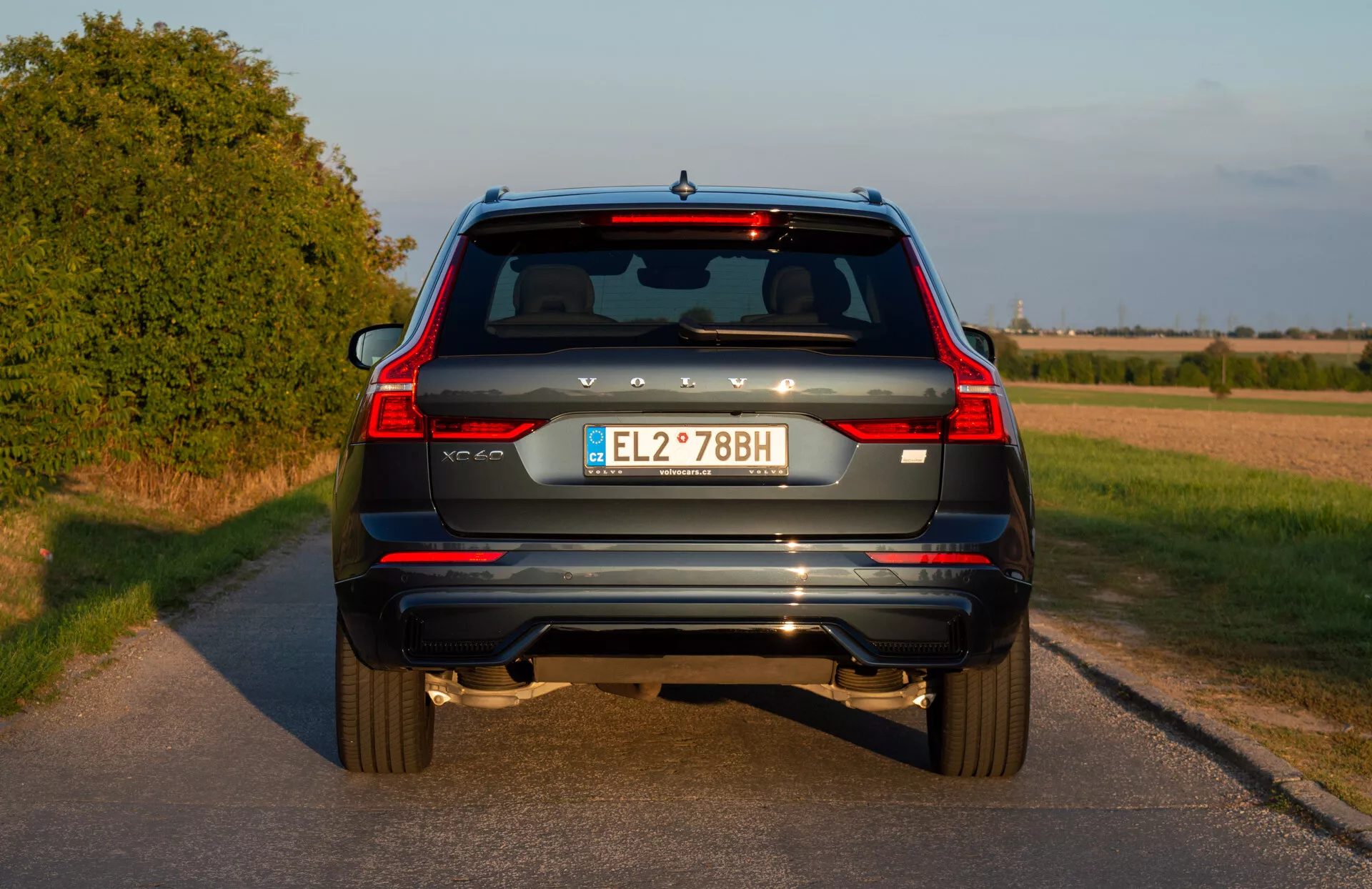 Tylny widok niebieskiego SUV Volvo XC60 Recharge Plug-In Hybrid z 2021 roku z pionowymi światłami tylnymi, podwójnymi końcówkami wydechu, reflektorami tylnymi, wycieraczką szyby tylnej, trzecim światłem stop, europejską tablicą rejestracyjną i kamerą cofania na wiejskiej drodze przed zachodem słońca