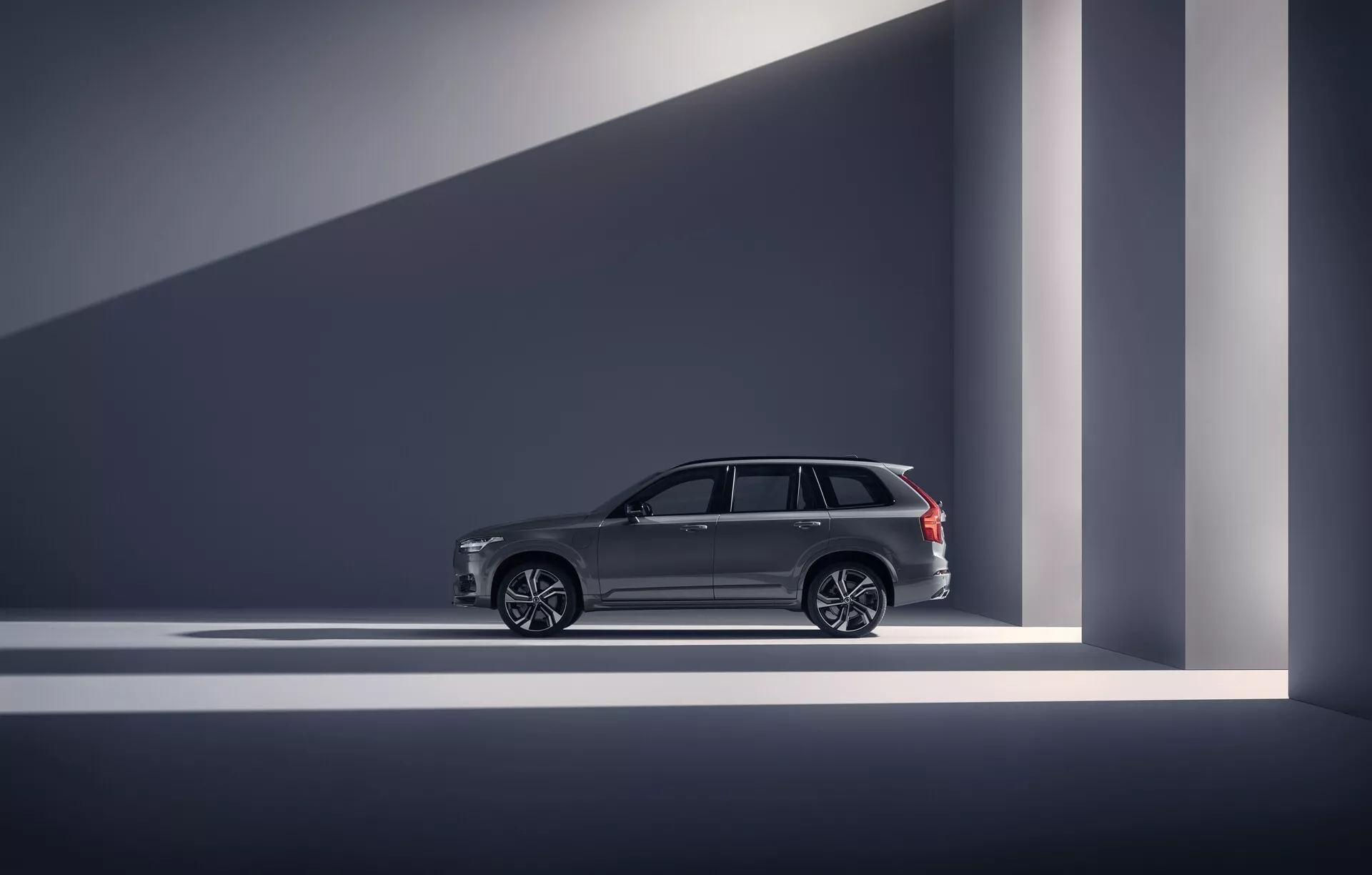 Profil boczny Volvo XC90 2015 (2. generacja) w kategorii luksusowych SUV-ów. Zdjęcie wykonane w zamkniętym studio lub salonie wystawowym, prezentujące linie projektowe pojazdu, eleganckie klamki drzwi, duże wieloramienne felgi aluminiowe oraz chromowane wykończenia okien. Relingi dachowe sugerują połączenie użyteczności i luksusu. Pojazd jest dobrze oświetlony dramatycznym oświetleniem, tworzącym geometryczne cienie, co uwypukla jego nowoczesny i ekskluzywny wygląd. Zaawansowane funkcje, takie jak bezkluczykowy dostęp, są sugerowane poprzez elementy dizajnerskie.
