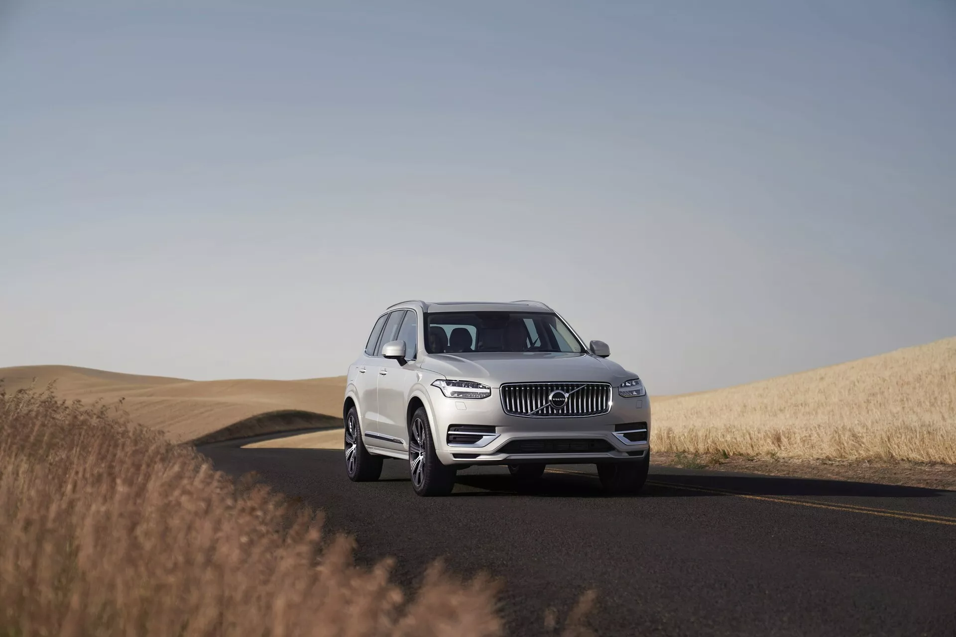 Białe SUV Volvo XC90 z 2015 roku na dwupasmowej drodze, uchwycone w świetle dziennym pod czystym niebem. Obraz pokazuje pojazd pod niskim do średniego kątem, podkreślając jego imponującą obecność i wymiary. Umieszczony na środku kadru z widokiem na trzy czwarte, zdjęcie uchwyca szczegóły projektu takie jak kratka wlotu powietrza, reflektory LED, emblemat i przedni zderzak, wskazując na zaawansowane systemy wspomagające kierowcę. Koła, felgi i ewentualnie rozmiar opon są wyraźnie widoczne. Spokojne otoczenie obejmuje drogę zakręcającą w prawo oraz złote pola pszenicy, podkreślając przydatność pojazdu do długich podróży w różnych krajobrazach.