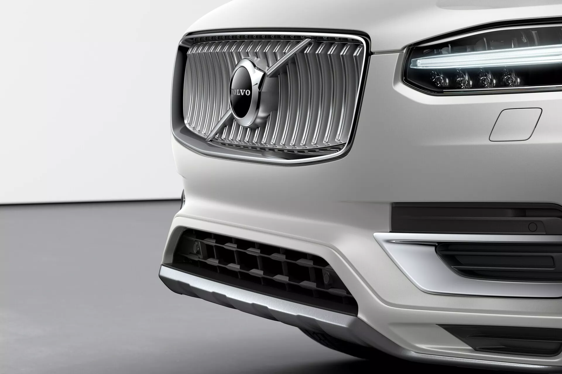 Zbliżenie przedniej części białego Volvo XC90 2015 drugiej generacji, podkreślające jego wyraźny grill z pionowymi przęsłami i charakterystycznym emblematem Volvo na środku. Chromowane wykończenie grilla kontrastuje z matową teksturą wokół niego. Widoczne są dzienne światła LED Młot Thora w zespolonych reflektorach. Dolny zderzak posiada duży wlot powietrza z srebrnym wykończeniem, co sugeruje efektywność aerodynamiczną i chłodzenie silnika. Zdjęcie zrobione z nieco niskiego kąta podkreśla elegancję, jakość wykonania oraz przedni, ekskluzywny design marki Volvo.