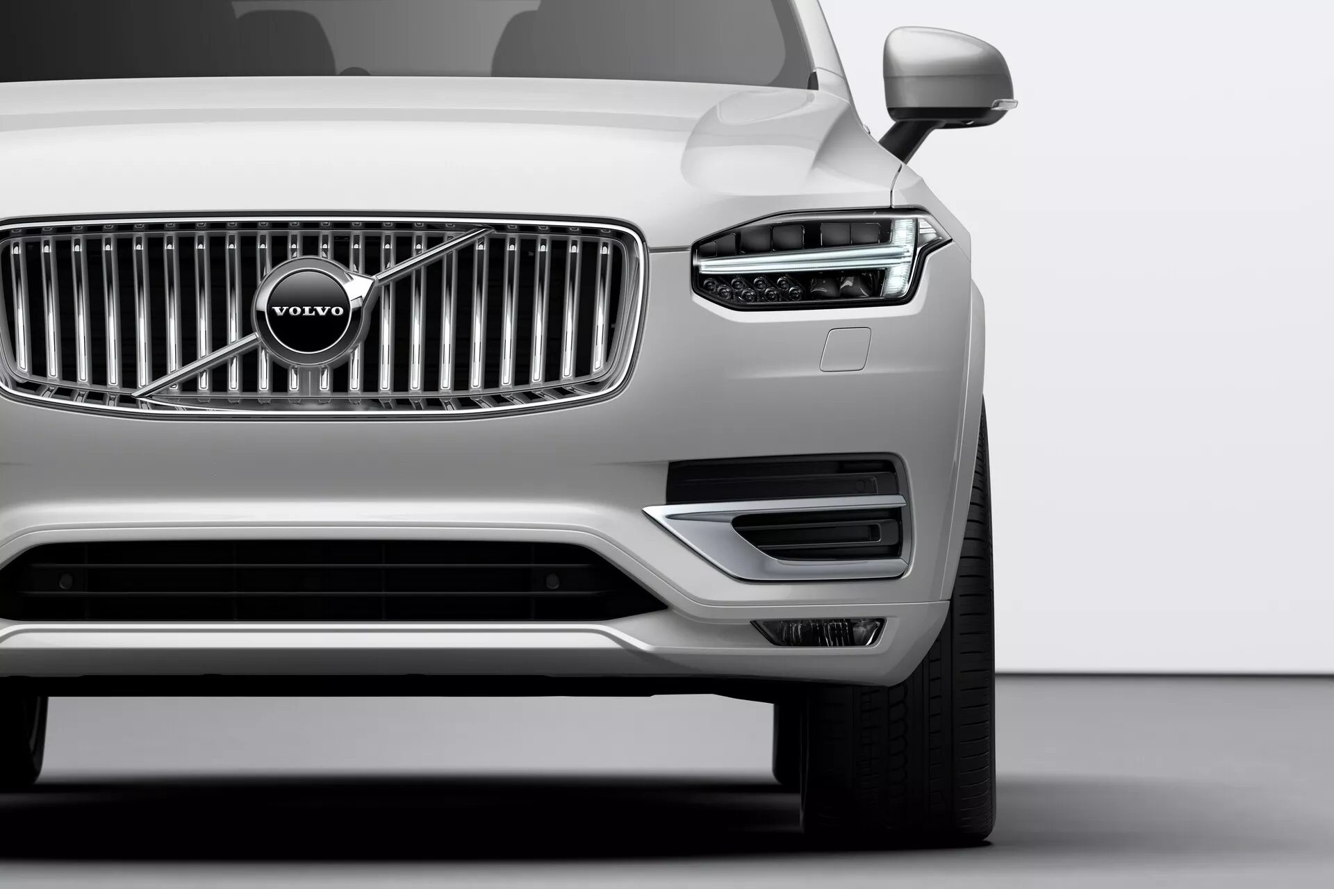 Zbliżenie przedniego kwartału srebrnego Volvo XC90 z 2015 roku (2. generacja) zrobione pod kątem niskim. Wyróżnia się widoczna chromowana krata z charakterystycznym emblematem Volvo, eleganckie reflektory z dziennymi światłami LED w kształcie litery T, zwane 'Młotem Thora', oraz dobrze zintegrowane czujniki parkowania w przednim zderzaku. Dolna część zderzaka zawiera wloty powietrza i obudowy lamp przeciwmgielnych otoczone srebrną listwą. Widoczne lusterka zewnętrzne sugerują zaawansowane funkcje technologiczne, takie jak kierunkowskazy lub systemy ostrzegania punktu martwego.