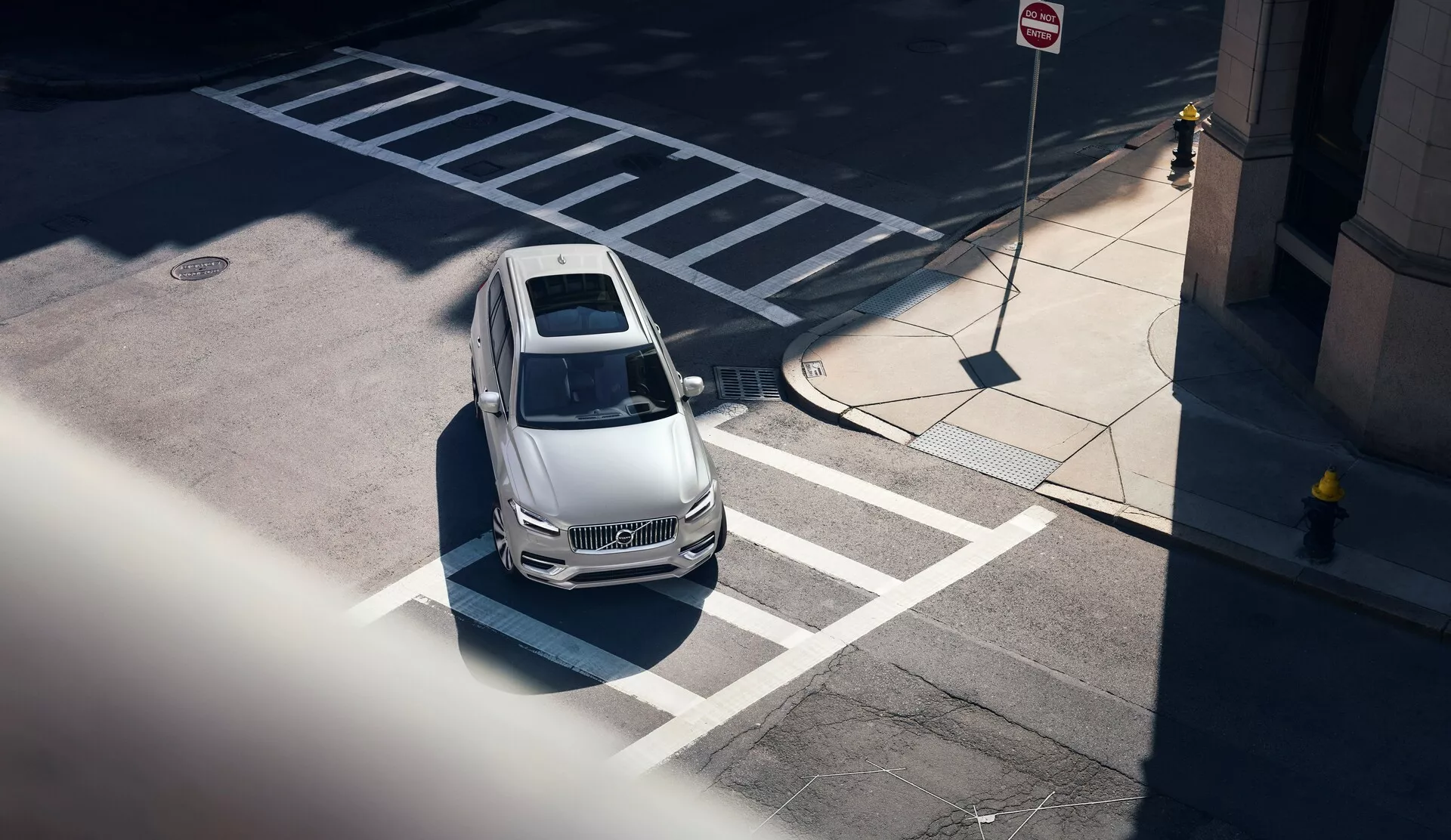 Widok z góry na Volvo XC90 2015 (2. generacja) z wysokiego kąta, widziane po przekątnej z lewego przedniego rogu; ukazujące jasny kaptur, dach i część strony kierowcy. Samochód, prawidłowo zaparkowany w wyznaczonym miejscu parkingowym blisko przejścia dla pieszych, ma kontury projektowe sugerujące aerodynamiczny styl, przeszkloną szybę odsłaniającą częściowo widoczne wnętrze, wyróżniający się dach panoramiczny i ostre linie boczne. Miejska sceneria jest podkreślona przez pobliskie znaki parkingowe i ruchowe, ze wyraźnymi cieniami sugerującymi oświetlenie w południe. Samochód również sugeruje nowoczesne funkcje technologiczne bez widocznych czujników czy kamer.