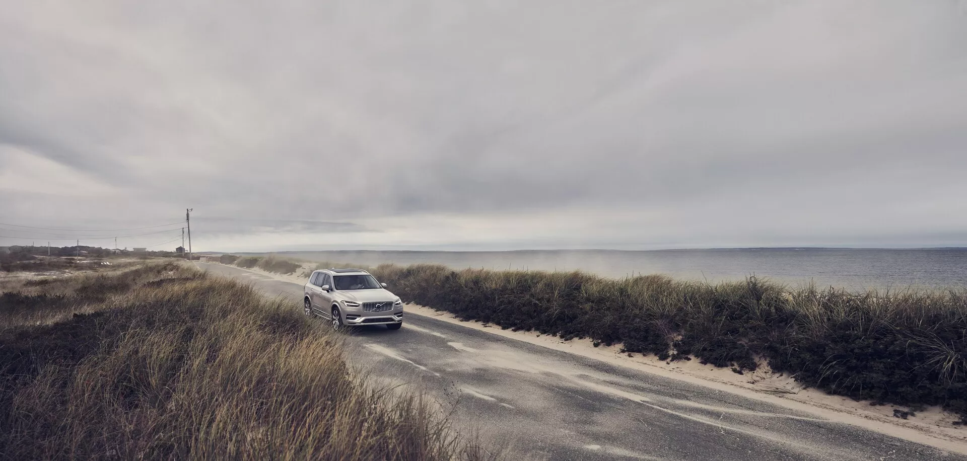 Szerokokątne zdjęcie Volvo XC90 z 2015 roku (2. generacja) jadącego wąską drogą nadmorską. Samochód zbliża się do kamery, podkreślając przedni grill, reflektory i zderzak. Na tle pochmurnego nieba i oceanu metaliczny, jasny kolor SUV-a wyróżnia się. Ta scena podkreśla połączenie luksusu i przygody pojazdu, sugerując zaawansowane funkcje technologiczne i bezpieczeństwa dla warunków jazdy nadmorskiej.
