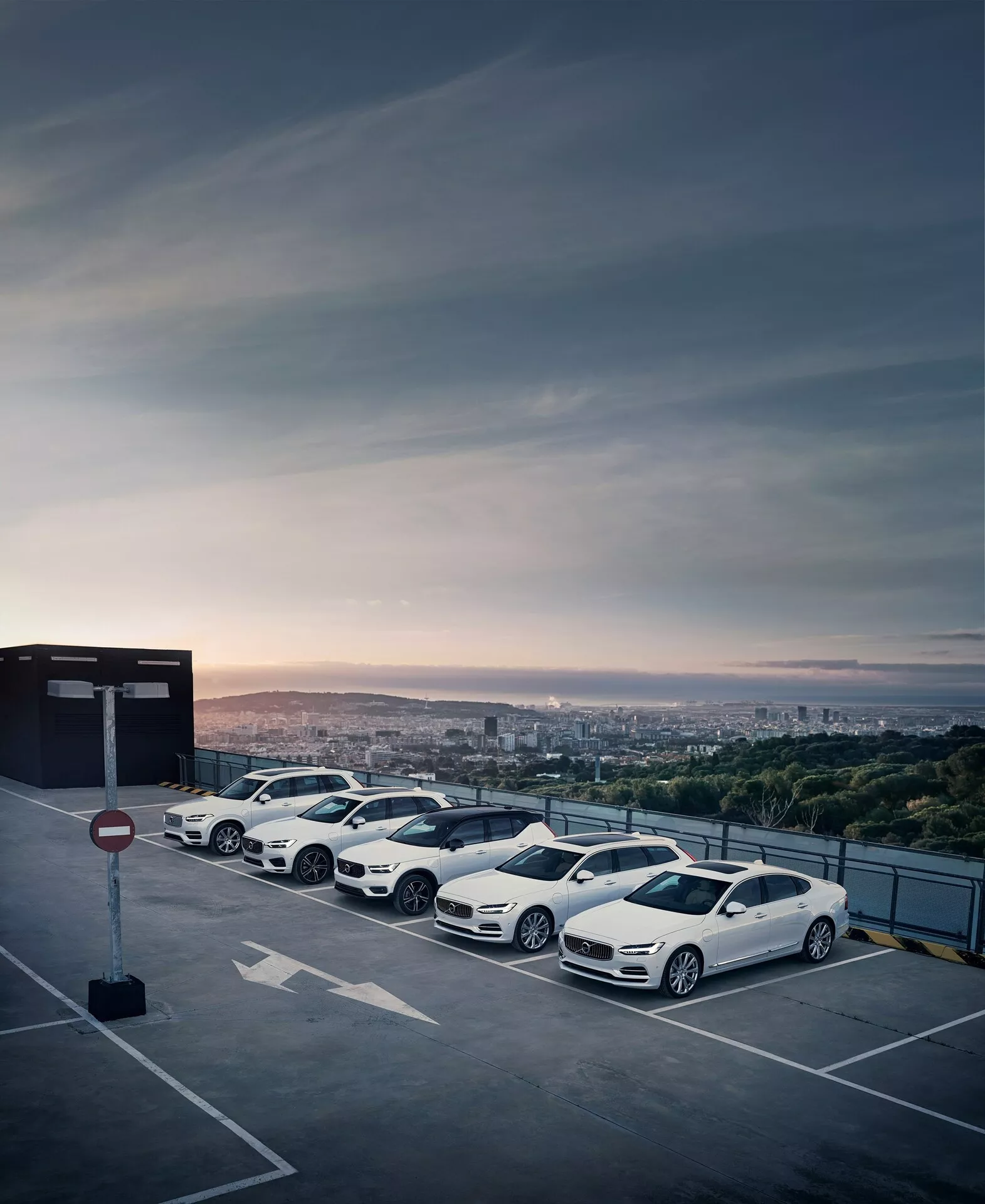 Linia czterech nowoczesnych, luksusowych samochodów Volvo XC90 2015 (2. generacja) zaparkowanych na najwyższym piętrze podwyższonej konstrukcji parkingowej podczas wschodu lub zachodu słońca z panoramą miasta w tle. Białe sedany mają aerodynamiczne wzory, wieloszprychowe felgi aluminiowe, aerodynamiczne lusterka boczne i przyciemniane szyby, ukazujące ich eleganckie kontury i dynamiczną estetykę.