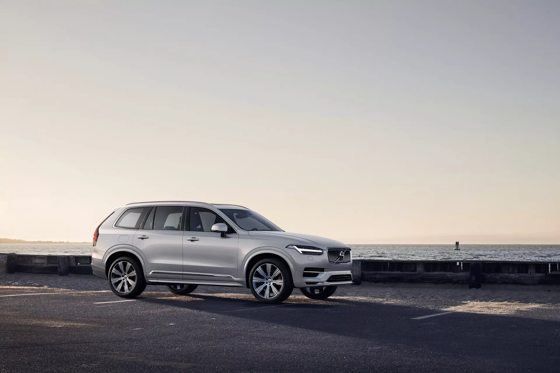 Profil boczny białego SUV-a Volvo XC90 2015 na nadmorskiej drodze, z widocznymi cechami designu, w tym wzorem kół, panelami drzwi, lusterkami bocznymi, relingami dachowymi i konfiguracją okien podkreślonymi miękkim światłem o świcie lub zmierzchu.