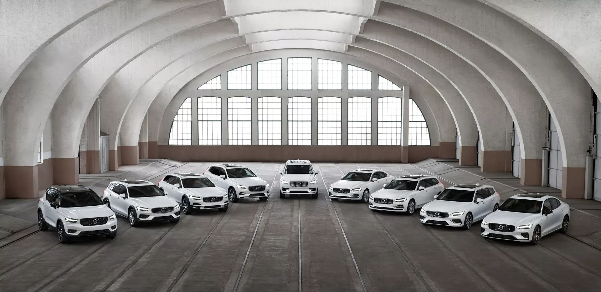 Linia ośmiu białych samochodów Volvo XC90 2015 drugiej generacji usytuowanych w przestronnym, architektonicznie klasycznym budynku z dużymi łukowatymi oknami i zakrzywionym sufitem. Pojazdy są skierowane w stronę aparatu pod różnymi kątami, prezentując swoje przednie części i cechy nadesignu, w tym reflektory LED, duże alufelgi i rzeźbione panele karoserii. Oświetlenie podkreśla elementy wzornictwa samochodów, a podłoga, oznaczona śladami opon, sugeruje używanie przestrzeni do przechowywania samochodów.