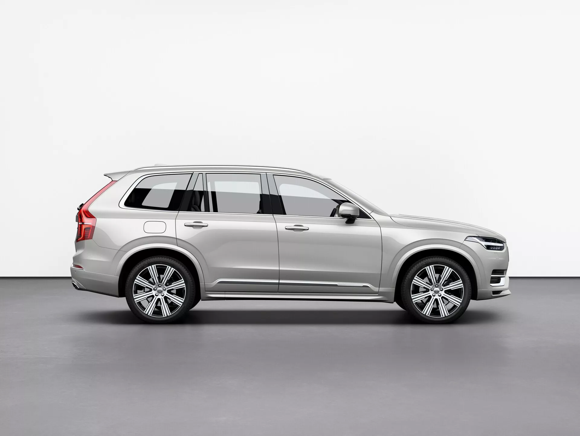 Boczny profil srebrnego SUV-a Volvo XC90 2015 (2. generacja), prezentujący jego felgi aluminiowe, relingi dachowe, lusterko boczne z zintegrowanym kierunkowskazem i szczegółowe linie nadwozia, umiejscowiony centralnie na prostym tle.