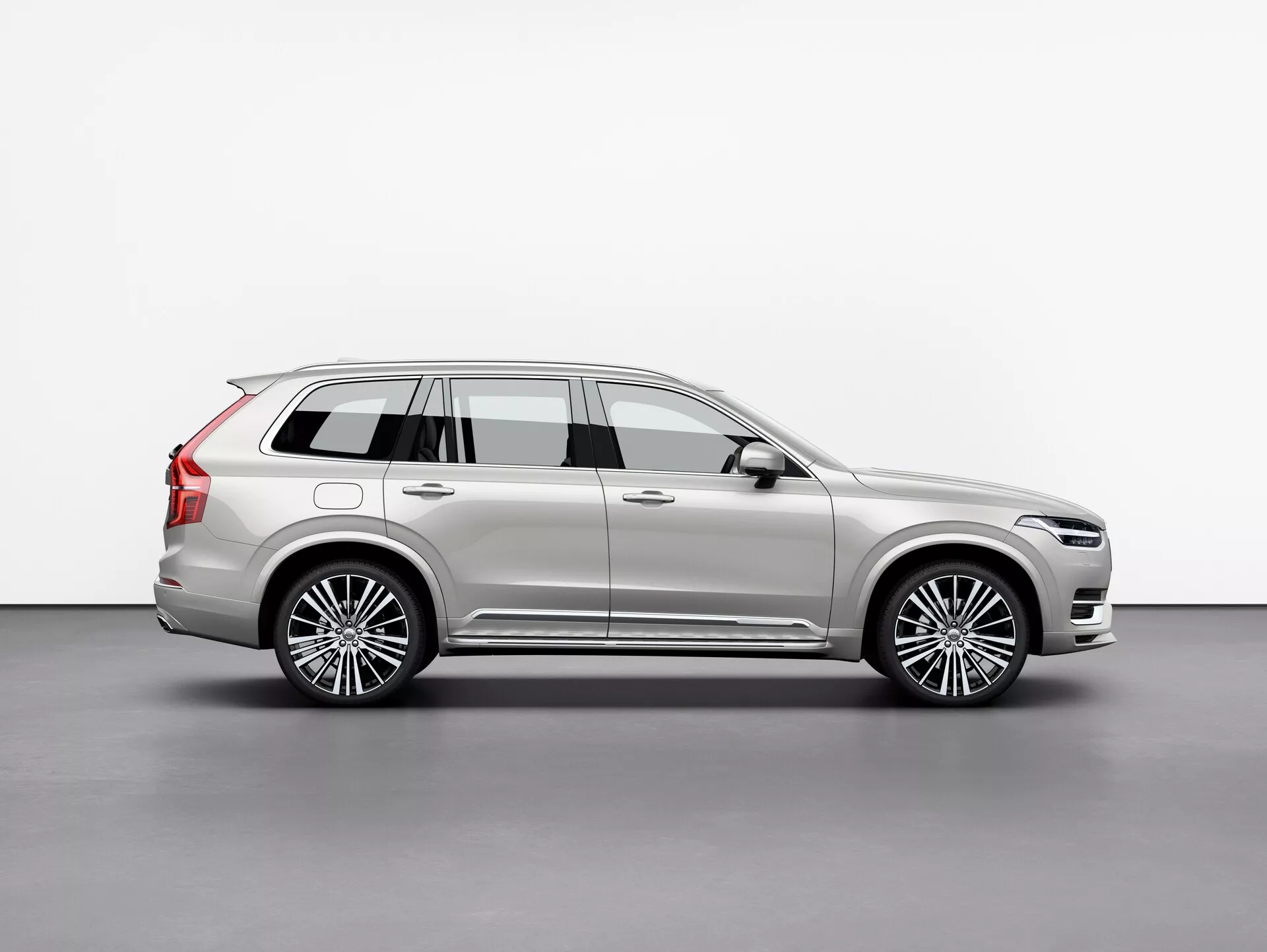 Profil boczny Volvo XC90 2015, luksusowego SUV-a w pełnym rozmiarze, uchwycony w studiu na białym tle. Obraz ukazuje długi rozstaw osi pojazdu, cztery drzwi, tylną klapę, wieloramienne felgi ze stopów metali, chromowane wykończenia, metaliczną powłokę lakierniczą i widoczne elementy projektowe, takie jak lusterka boczne i relingi dachowe. Starannie oświetlona scena podkreśla linie i krzywizny karoserii, odzwierciedlając jej luksusowy charakter. Pojazd jest ustawiony w taki sposób, aby zapewnić wyraźny widok na jego całkowitą długość, wysokość i posturę, co świadczy o SUV-ie z trzema rzędami siedzeń i dużą przestrzenią bagażową.