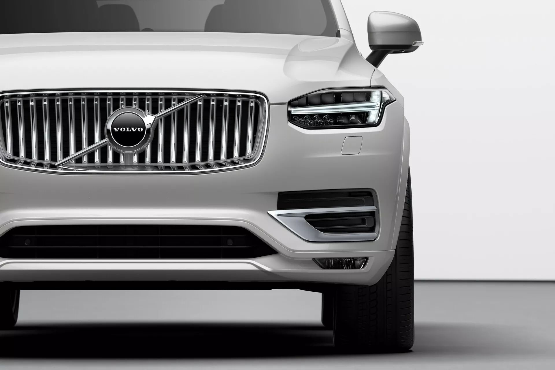 Nisko ustawione zdjęcie lewego przedniego rogu srebrnego Volvo XC90 (2. generacja) z 2015 roku w studio, pokazujące charakterystyczny wzór LED w kształcie młota Thora na reflektorze, pionowe chromowane listwy grilla z emblematem Volvo, minimalistyczny przedni zderzak z wlotami powietrza, lewe lusterko boczne, wieloramienne aluminiowe koło i nieskazitelne metaliczne wykończenie, obrazując zaawansowaną technologię oświetlenia, styl marki i cechy aerodynamiczne.