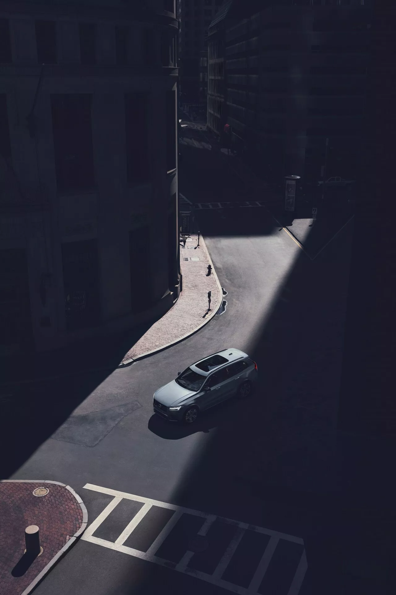Widok z lotu ptaka na sedana Volvo XC90 2015 (2. generacji) zaparkowanego na zacienionej miejskiej ulicy, uchwycony z dużej wysokości pod kątem ukośnym. Wyraźnie widoczne są bagażnik, dach i tylna szyba samochodu, z tylną częścią pojazdu oświetloną światłem słonecznym. Obraz pokazuje tylną klapę bagażnika, kształt tylnej szyby i górną część bagażnika, podczas gdy cienie ukrywają specyficzne detale designu, takie jak tylne światła i zderzak. Widoczna jest gładka, aerodynamiczna linia dachu, bez widocznych bagażników dachowych lub szyberdachów. Samochód jest zaparkowany przy krawężniku w pobliżu fasad budynków i przejścia dla pieszych, co podkreśla izolację pojazdu w zacienionym miejskim otoczeniu.