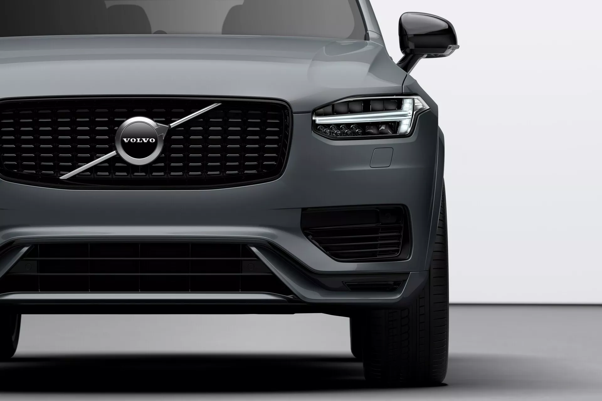 Częściowy, asymetryczny przód Volvo XC90 2015 (2. generacja) pokazujący nowoczesne elementy designu jak wyraźne logo Volvo na grillu, LED-owe światła dzienne 'Młot Thora', aerodynamiczny zderzak, eleganckie lusterko boczne z wbudowanym kierunkowskazem oraz ciemny lakier w neutralnym szarym świetle.