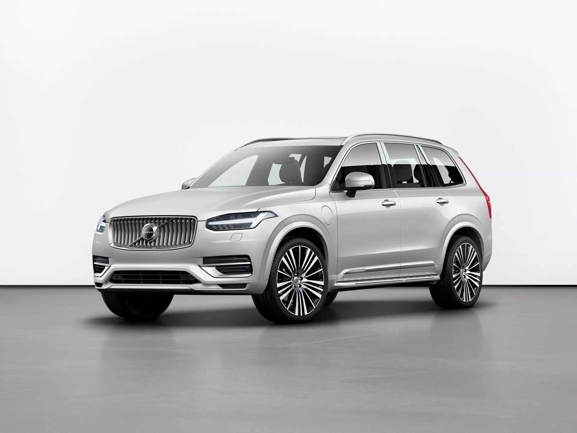 Zdjęcie studyjne luksusowego SUV-a Volvo XC90 2015 (2. generacja), przedstawione pod kątem przednim trzy czwarte, ukazujące przedni grill z charakterystycznym logo, eleganckie reflektory LED, masywny przedni zderzak i długi rozstaw osi z dużymi felgami aluminiowymi. Lusterka boczne znajdują się poniżej słupka A, z oknami wykończonymi chromowanymi listwami i relingami dachowymi. Neutralny kolor lakieru emanuje elegancją, a zdjęcie podkreśla linie i kontury samochodu z równomiernym oświetleniem na jednolitym tle. Prawdopodobne cechy obejmują technologie wspomagania kierowcy i zaawansowane systemy informacyjno-rozrywkowe.