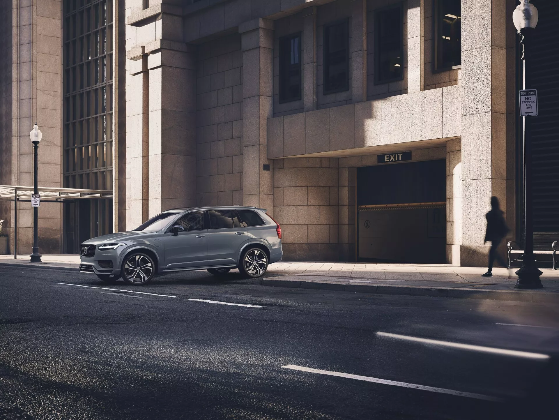 Nowoczesny SUV Volvo XC90 2015 (2. generacja) zaparkowany na miejskiej ulicy, uchwycony z perspektywicznego kąta, pokazujący profil boczny oraz część przedniego grilla. Błyszczący metaliczny lakier samochodu podkreśla jego dobry stan, wieloramienne felgi aluminiowe i przyciemniane szyby. Zdjęcie zrobione z poziomu oczu przeciętnego dorosłego, ukazujące auto na pierwszym planie, otoczone miejskimi elementami, takimi jak fasada budynku, sylwetka pieszego i latarnia uliczna w porannym lub późno popołudniowym świetle słonecznym.