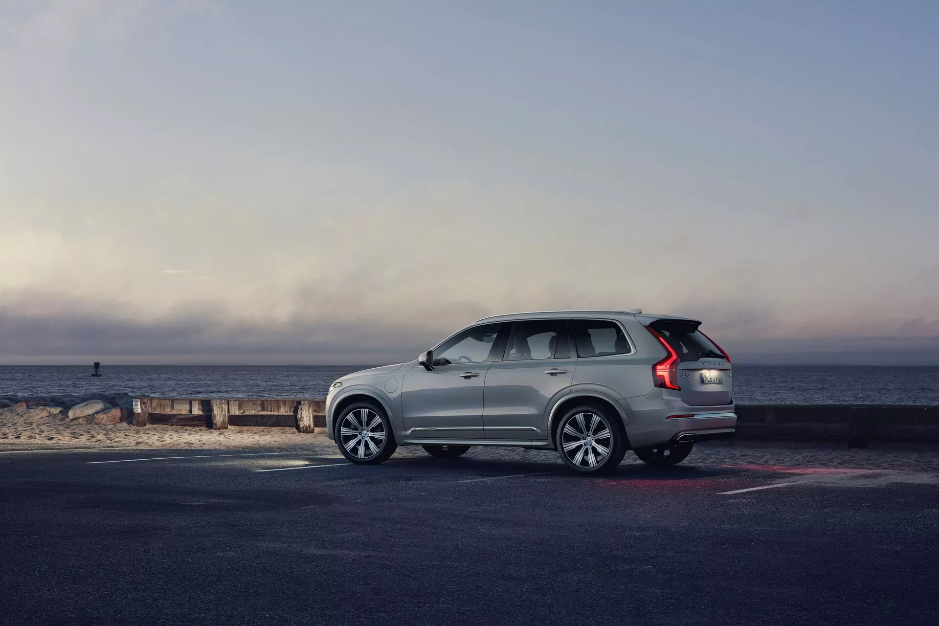Widok z profilu SUV-a Volvo XC90 z 2015 roku (2. generacja) zaparkowanego przy brzegu, z pojazdem skierowanym w prawo. Kąt ukazuje bok i tył, ukazując elementy stylistyczne, takie jak wieloramienne felgi aluminiowe, wydłużone tylne światła LED, relingi dachowe, wznoszącą się ku tyłowi linię szyb oraz wpuszczane klamki drzwi. Jasny kolor pojazdu wyróżnia się na tle ciemniejszego chodnika i plaży, oświetlonych naturalnym światłem porannym lub późnym popołudniowym, z malowniczym tłem horyzontu, gdzie niebo spotyka się z morzem. Cała kompozycja wywołuje wrażenie przygody, luksusu i wolności.