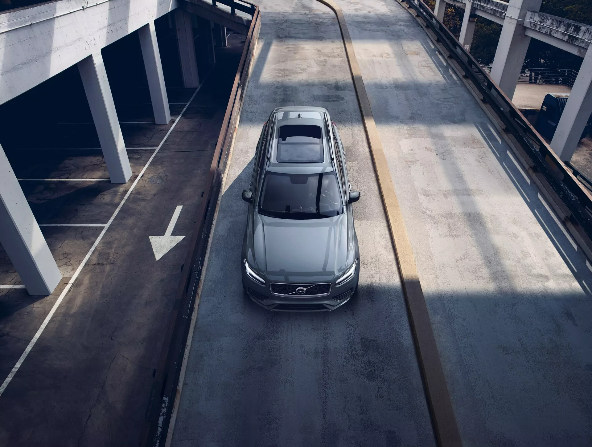 Widok z lotu ptaka na szary lub srebrny sedan Volvo XC90 2015 poruszający się po wielopoziomowej drodze miejskiej, ukazujący panoramiczny dach, aerodynamiczny design, eleganckie reflektory LED i duże felgi aluminiowe, sugerując nowoczesny i luksusowy pojazd.