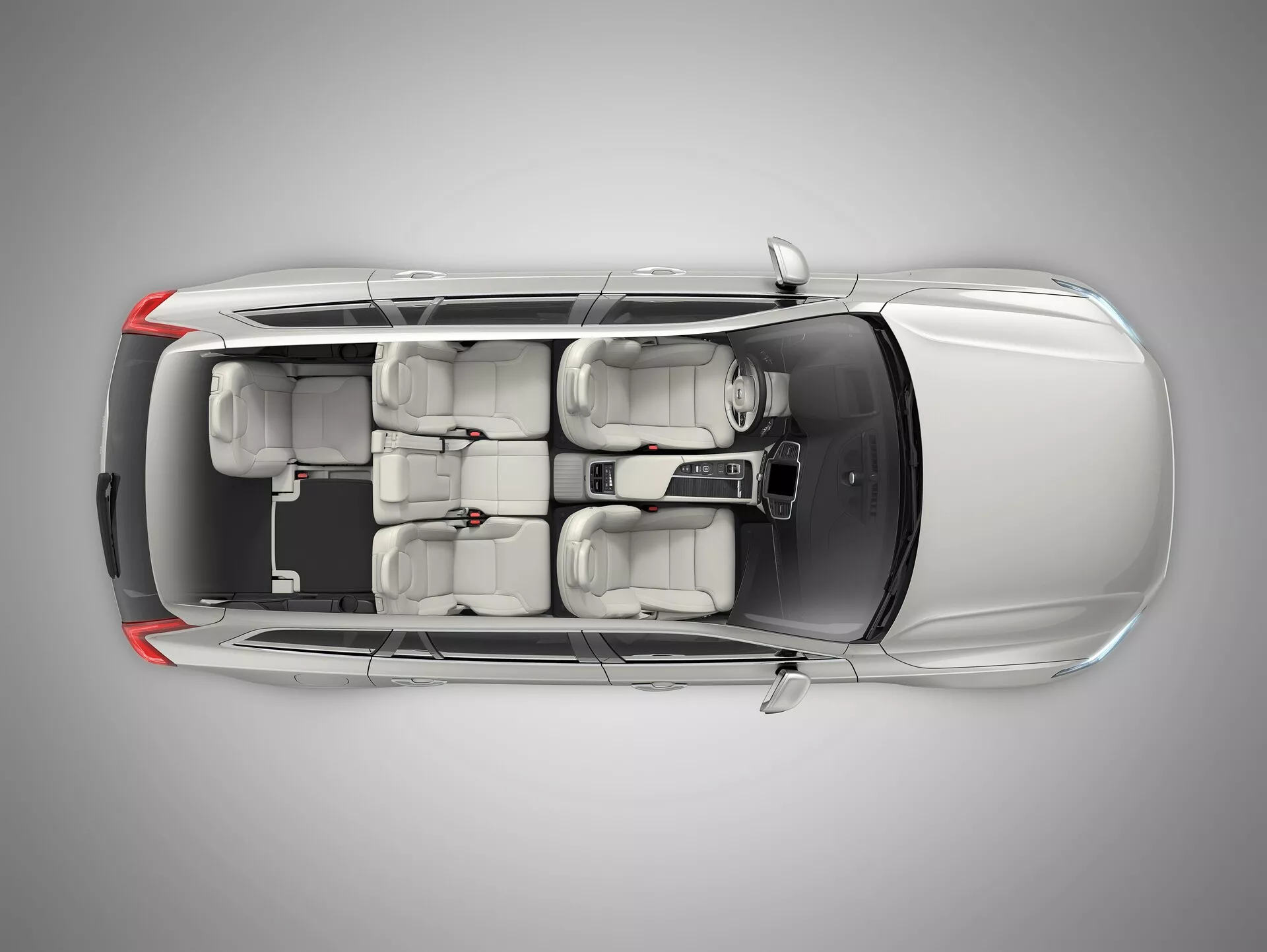 Widok z lotu ptaka na dobrze utrzymanego srebrnego sedana Volvo XC90 drugiej generacji z 2015 roku. Obraz uchwyca całą długość i szerokość auta, pokazując cały dach, nadwozie i otoczenie. Wszystkie okna i drzwi są zamknięte, klamki drzwi są zintegrowane, a boczne lusterka cofnięte. Dach nie ma szyberdachu, a warstwa farby jest równomiernie odbijająca światło. Wnętrze jest widoczne przez przejrzysty dach, ukazując beżową lub jasnoszarą tapicerkę, dwa rzędy siedzeń z zagłówkami i standardowe opony. Kształty obudowy reflektorów przednich i tylnych są widoczne, lecz mało szczegółowe.