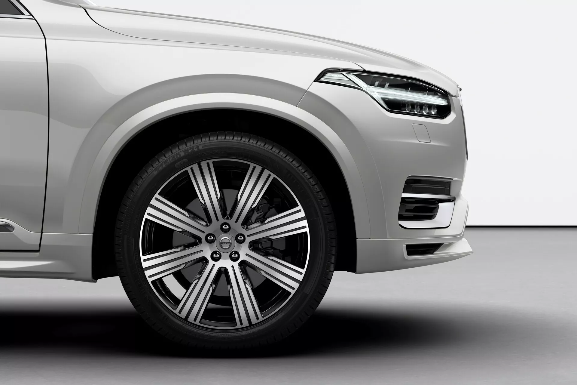 Zbliżenie, niskokontowy zrzut przedniego panelu Volvo XC90 2015 2. generacji, z przednim kołem po stronie kierowcy, wieloszprychowym aluplastowym designem, systemem hamulców tarczowych, widocznym zaciskiem hamulca i oponą drogową. Obraz ukazuje przedni zderzak z dużym wlotem powietrza, jasną metaliczną farbą i gładkimi zakrzywionymi liniami aerodynamicznymi.