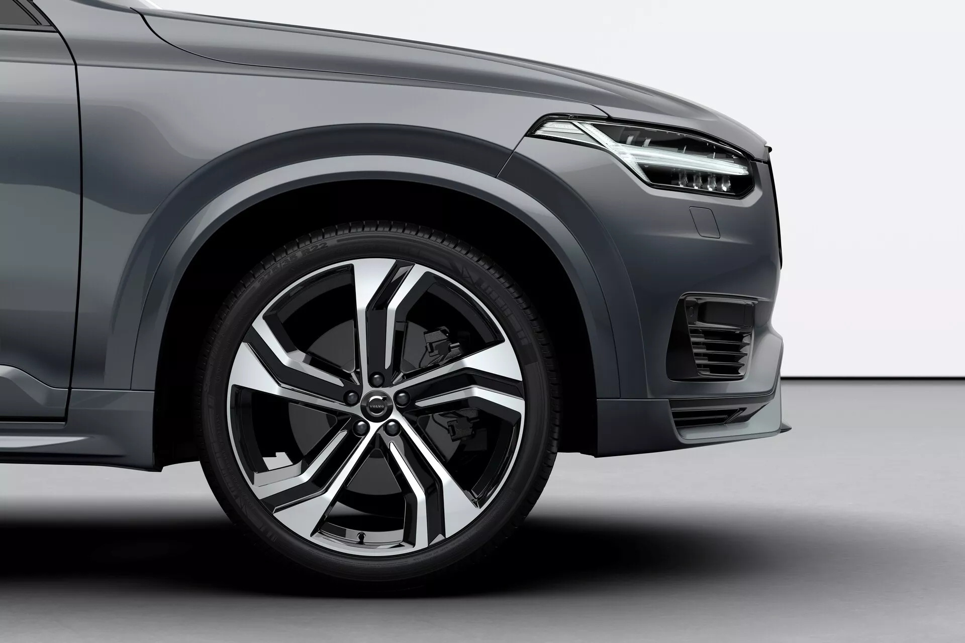 Zbliżenie z niskiego kąta na przednie koło i dolną część Volvo XC90 2015 (2. generacja). Zdjęcie przedstawia wieloszprychowe koło aluminiowe z wykończeniem w kolorze srebrnym i czarnym, oponę niskoprofilową skoncentrowaną na wydajności na drodze oraz częściowo widoczne eleganckie, poziome reflektory. Obraz ukazuje również wyprofilowany przedni zderzak z małą sekcją kratki, sugerując luksusowy, nowoczesny SUV. Samochód zajmuje większość kadru, a tło jest neutralne, prawdopodobnie studyjne.