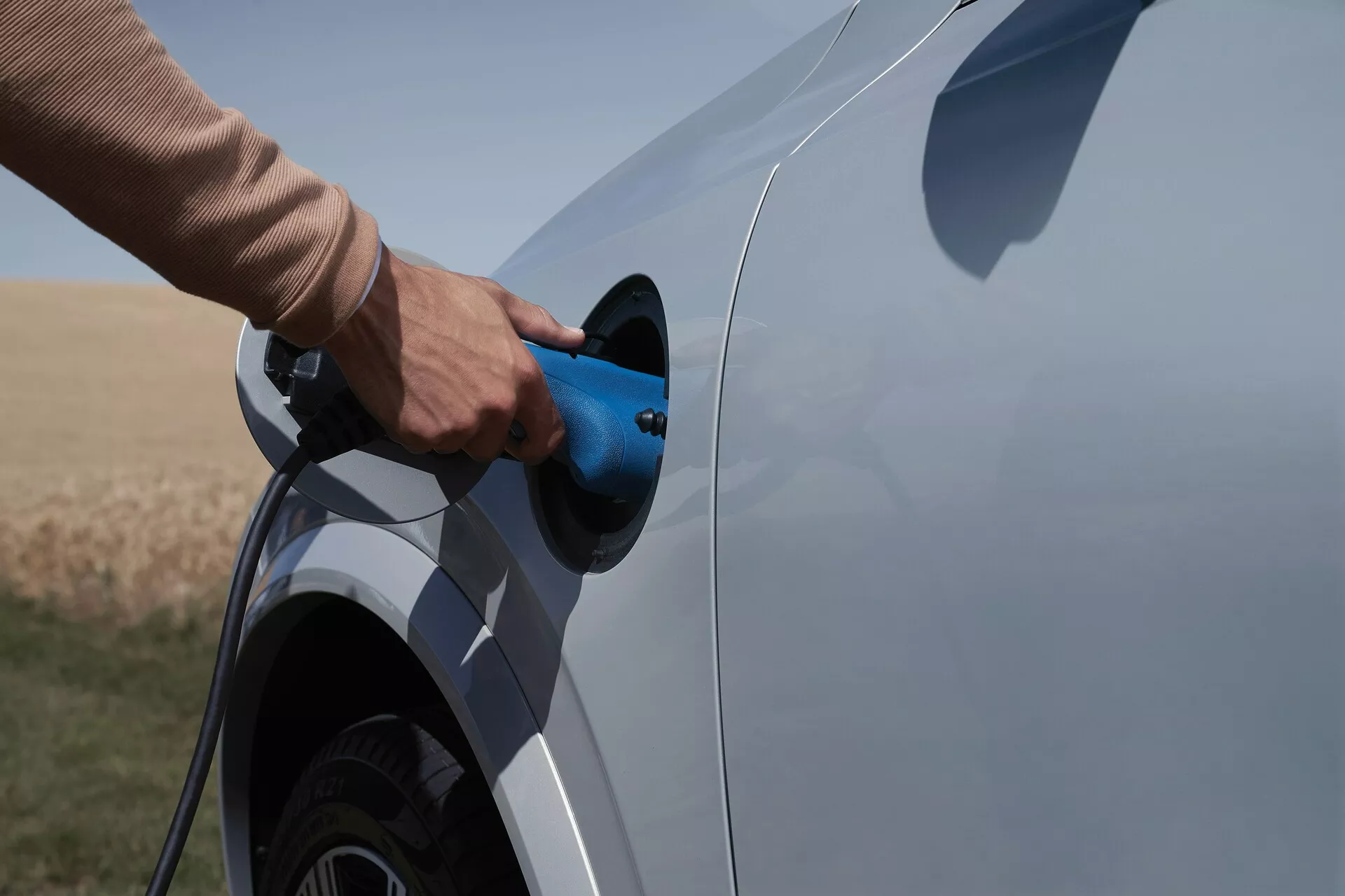 Widok z bocznego kąta na elektryczny lub hybrydowy plug-in pojazd Volvo XC90 2015 drugiej generacji podczas procesu ładowania. Zdj�cie uchwyca bok pojazdu z naciskiem na port i wtyczkę ładowania, częściowo pokazując tylne koło i segment dolnej części samochodu. Niebieski kabel ładowania wkładany, tło otwartego obszaru naturalnego, wskazuje na wiejskie otoczenie.