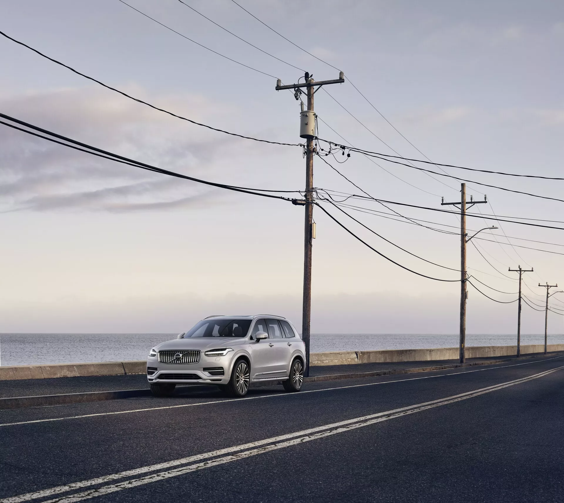 Volvo XC90 drugiej generacji z 2015 roku, luksusowe SUV zaparkowane na asfaltowej drodze równoległej do linii brzegowej oceanu, sfotografowane w świetle dziennym przy miękkim rozproszonym świetle i długich cieniach. Widok z bocznego przedniego kąta, podkreślający eleganckie elementy designu, w tym reflektory, grille i kontury maski, kolor jasnosrebrny lub blady szampański kontrastujący z ciemną drogą i naturalnym tłem.