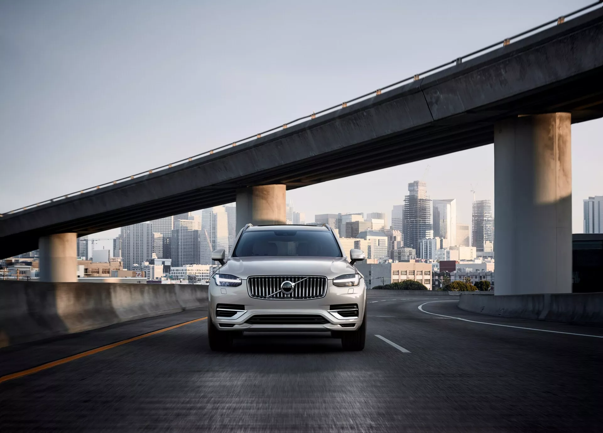 Frontalny kadr drugiej generacji SUV-a Volvo XC90 z 2015 roku na pustym wiadukcie, z panoramą miasta w tle. Kąt kamery jest niski, podkreślając przedni grill, reflektory LED, chromowane akcenty, felgi aluminiowe i zderzak z czujnikami, wskazujące na asystenta parkowania lub system unikania kolizji. Perłowy lub metaliczny lakier samochodu błyszczy w świetle dziennym, podkreślając jego luksus i nowoczesny design w miejskim otoczeniu.