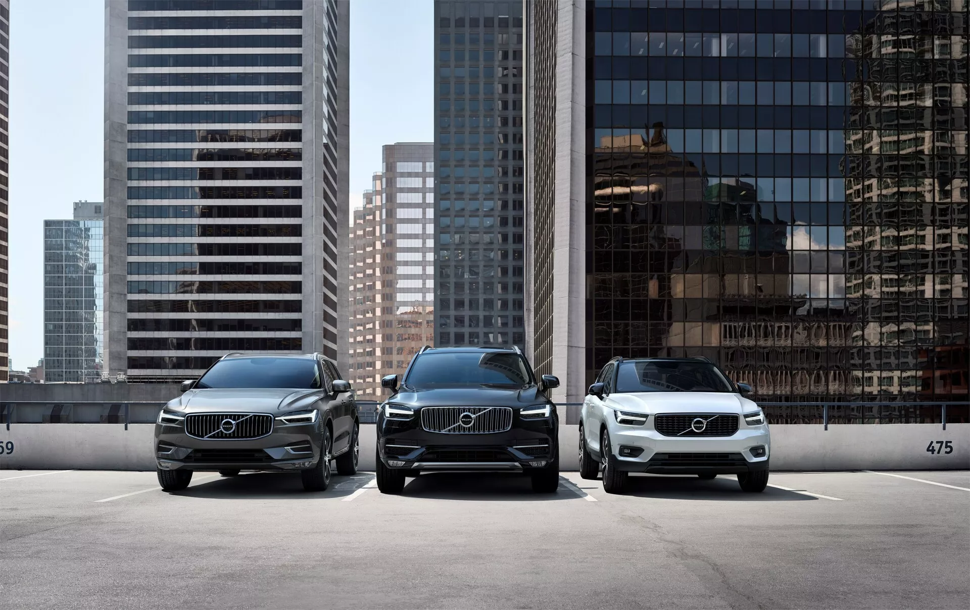 Trzy luksusowe samochody Volvo XC90 2015 (2. generacja) zaparkowane równolegle, skierowane przed kamerę, z nowoczesnym miejskim krajobrazem w tle. Frontalna perspektywa pojazdów ukazuje eleganckie reflektory LED, duże grille z wyraźnymi emblemami Volvo i czysty, elegancki design. Samochód na pierwszym planie po prawej stronie jest jaśniejszy, co podkreśla różnorodność dostępnych kolorów. Są zaparkowane za krawężnikiem z widocznym numerem 475, sugerującym wyznaczoną strefę parkowania lub układ wystawowy w salonie samochodowym.