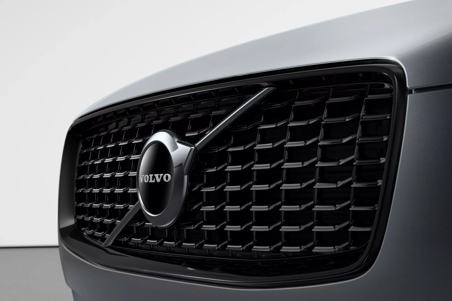 Zbliżenie przedniej części Volvo XC90 z 2015 roku (2. generacja) koncentrujące się na osłonie chłodnicy, zawierające gęsty wzór z siatki z prostokątnymi elementami, chromowanym emblematem Volvo na środku z strzałką skierowaną ukośnie w górę w prawo i widokiem na ciemną, elegancką karoserię samochodu po lewej stronie. Obrazek podkreśla estetykę projektu i rozpoznawalność marki.