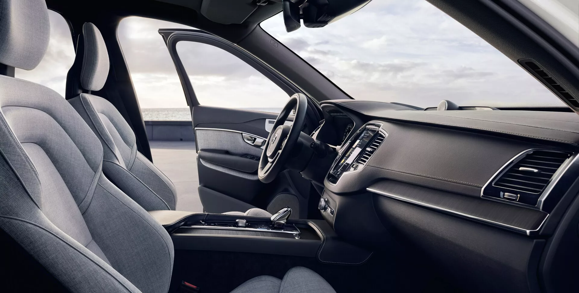 Ten obrazek przedstawia wnętrze drugiej generacji Volvo XC90 z 2015 roku z perspektywy kierowcy. Nieco podwyższony kąt umożliwia dokładny widok przednich siedzeń i deski rozdzielczej, z lewym fotelem kierowcy obitym teksturowaną tkaniną, z widocznym zagłówkiem i wsparciem bocznym. Wielofunkcyjna kierownica ma kilka przycisków sterowania, a elegancka deska rozdzielcza zawiera symetryczne otwory wentylacyjne i częściowo widoczny centralny ekran infotainment. Konsola centralna pokazuje dźwignię automatycznej skrzyni biegów i inne funkcje sterujące, podkreślając ergonomiczną i praktyczną konstrukcję.