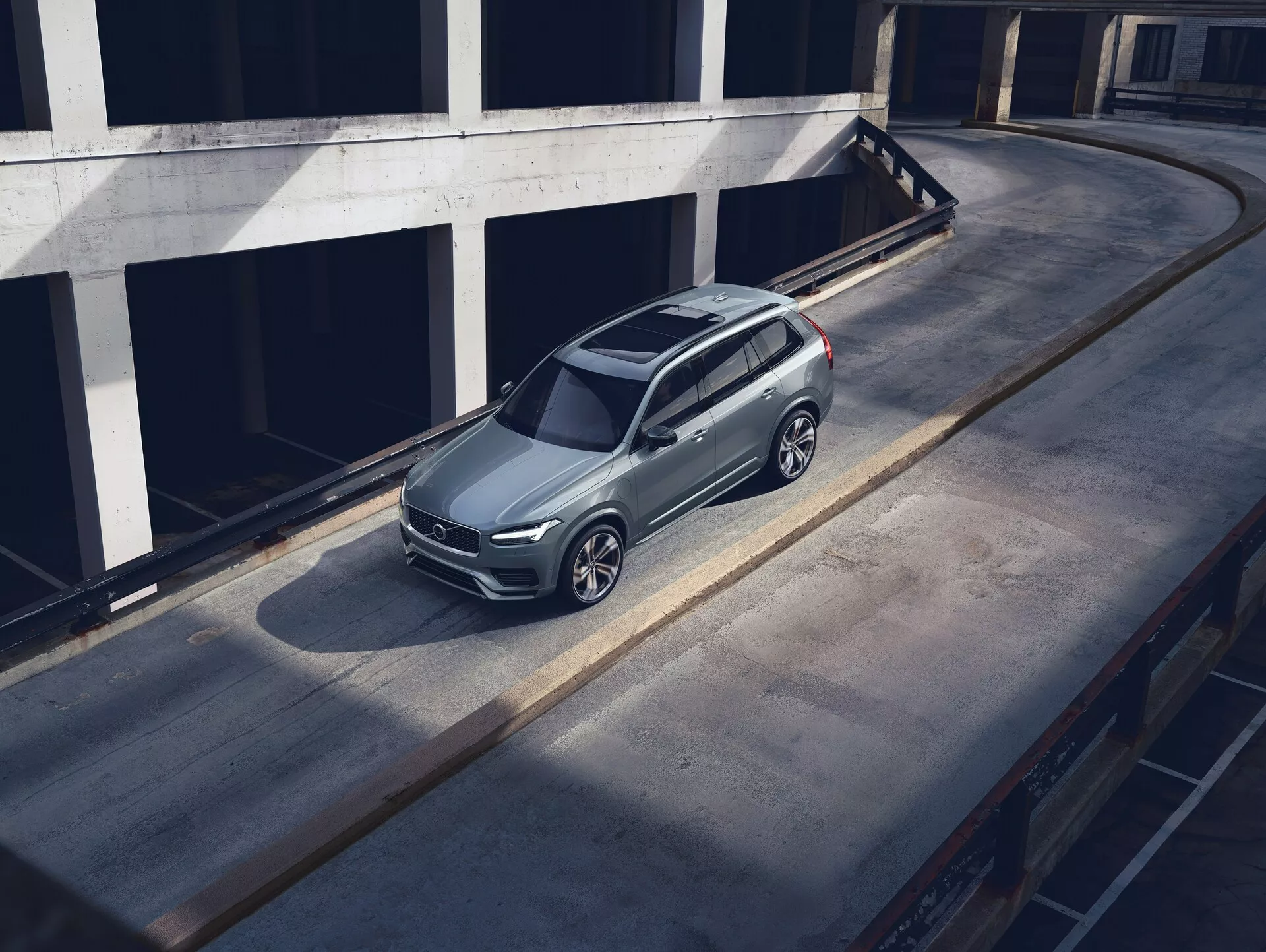 Widok z lotu ptaka na zaparkowanego SUV-a Volvo XC90 z 2015 roku na betonowej powierzchni, prezentujący jego elegancki, nowoczesny design z metalicznym wykończeniem lakieru, charakterystycznym przednim grillem, widocznymi reflektorami i skomplikowanymi alufelgami. Zdjęcie podkreśla dach, maskę i lewą stronę pojazdu, a cień wskazuje na poranne lub późnopopołudniowe światło. Otoczenie wygląda na górny poziom wielopoziomowej konstrukcji parkingowej, co zwiększa znaczenie pojazdu w czystym, miejskim środowisku.