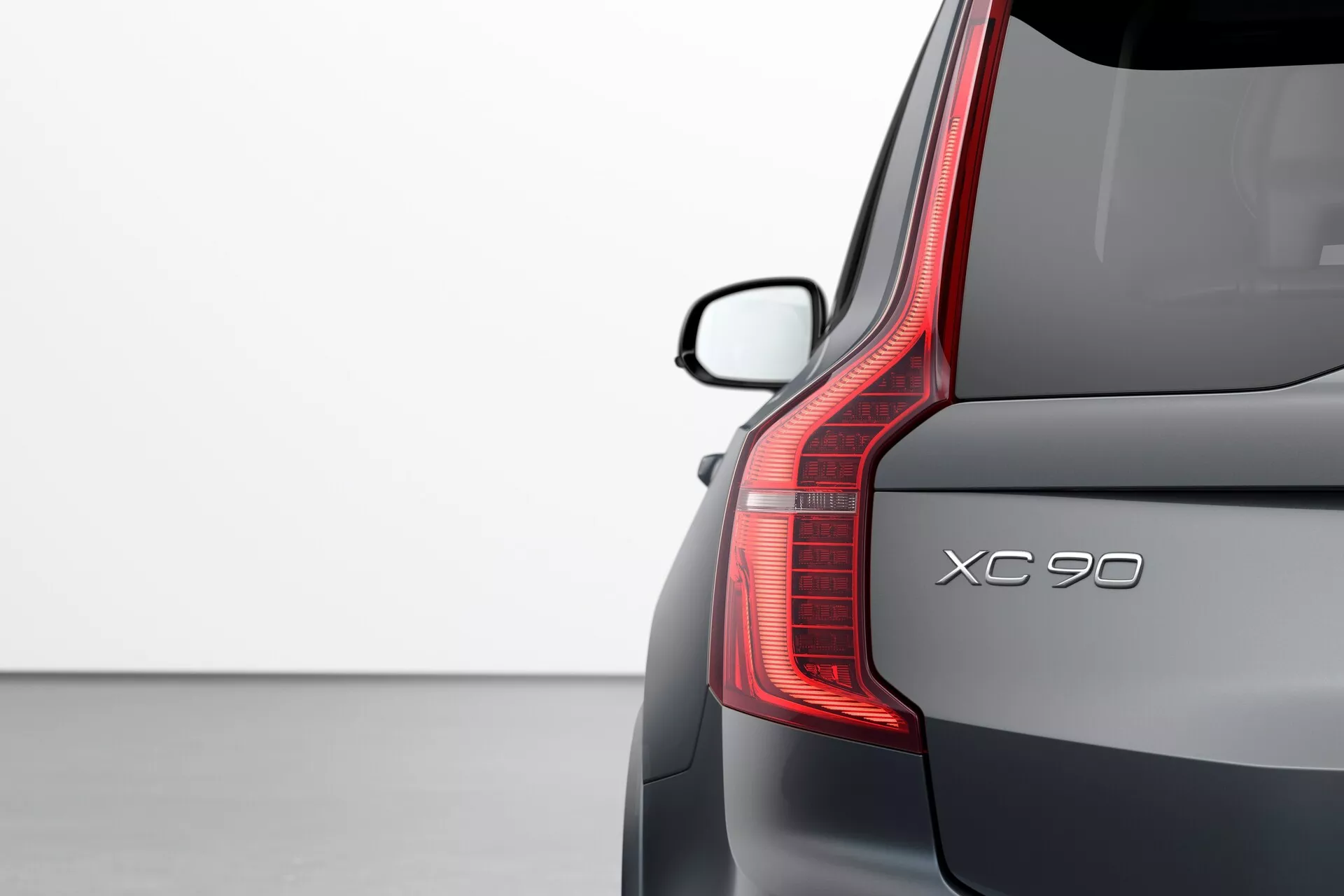 Zbliżenie tylnego rogu Volvo XC90 2015 (2. generacji) podkreślające szczegóły wzornictwa: pionowe tylne światło LED, nazwa modelu 'XC90', elegancka tylna szyba ze spojlerem oraz wykończenie premium metalicznym lakierem na jasnym tle.