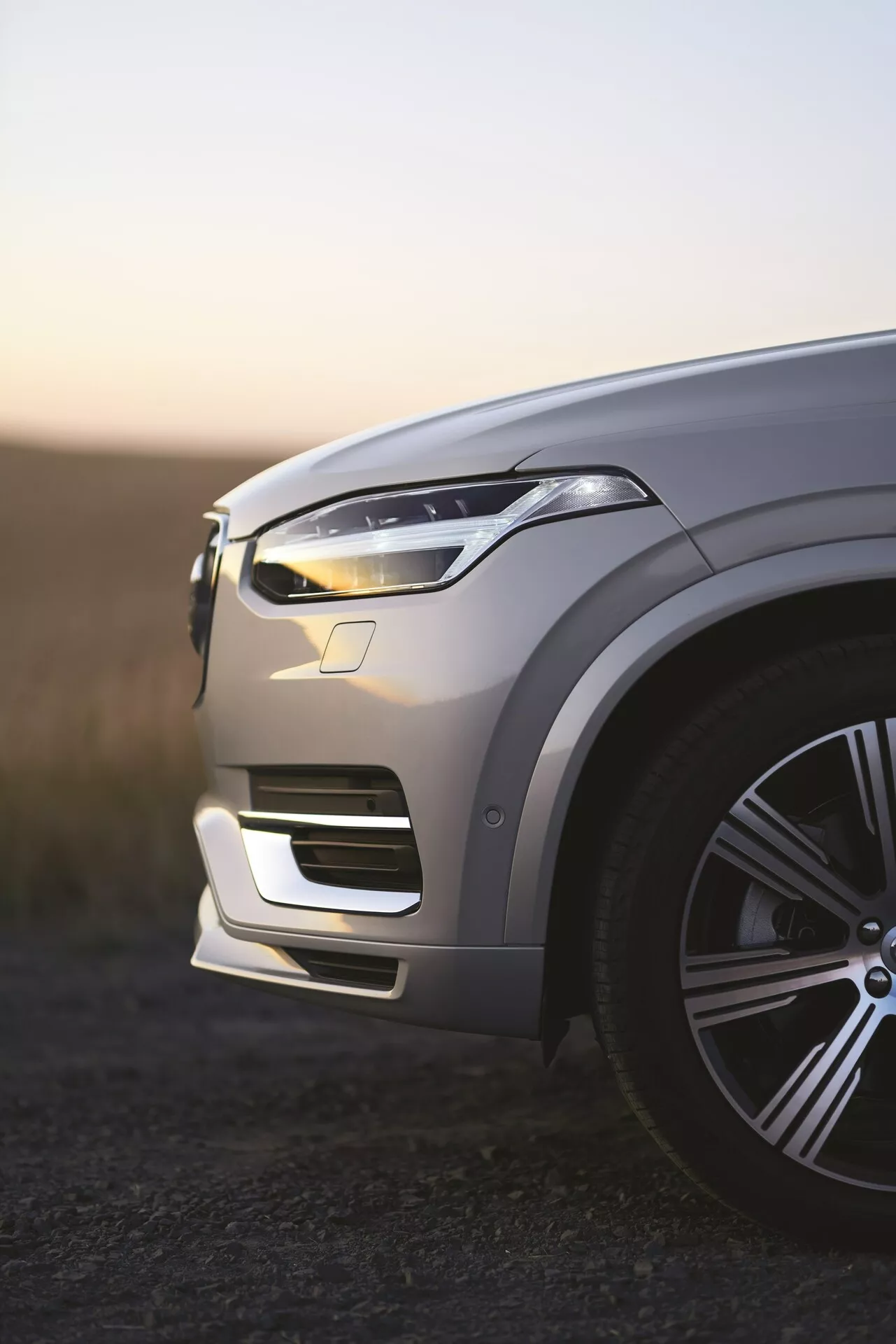 Widok z lewej przedniej ćwiartki luksusowego SUV-a Volvo XC90 2. generacji z 2015 roku o świcie, podświetlane światła dzienne LED, metaliczny lakier odbijający światło otoczenia, stylowe duże felgi aluminiowe, złożone kontury maski, elegancki design przednich świateł LED, zintegrowany przedni zderzak z wlotami powietrza, widoczne potencjalne czujniki wspomagania kierowcy, płytka głębia ostrości z rozmytym tłem.
