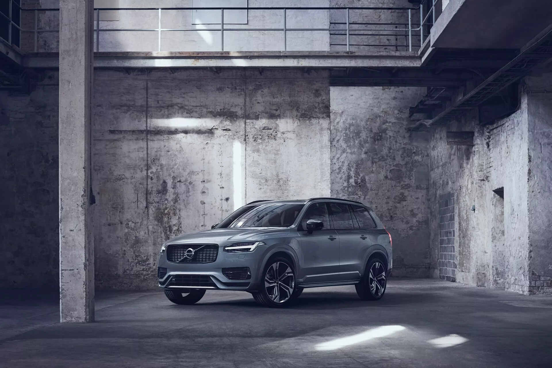Obraz metalicznie koloryzowanego Volvo XC90 2015 zaparkowanego w słabo oświetlonym garażu w stylu industrialnym. Uchwycony z przedniego widoku trzech czwartych, pokazując bok kierowcy z wyraźnie widocznym przodem i profilem bocznym. Samochód posiada eleganckie reflektory LED, dużą przednią kratkę, prawdopodobnie czujniki parkowania oraz duże aluminiowe felgi z wieloramiennym wzorem. Lusterka boczne są aerodynamiczne i mogą zawierać zaawansowane funkcje technologiczne, takie jak wskaźniki sygnalizacji i systemy ostrzegawcze martwego pola. Obraz podkreśla luksusowy, ale użyteczny design SUV-a z dużym prześwitem i potencjalnymi funkcjami zamontowanymi na dachu, takimi jak dach słoneczny lub panoramiczny.