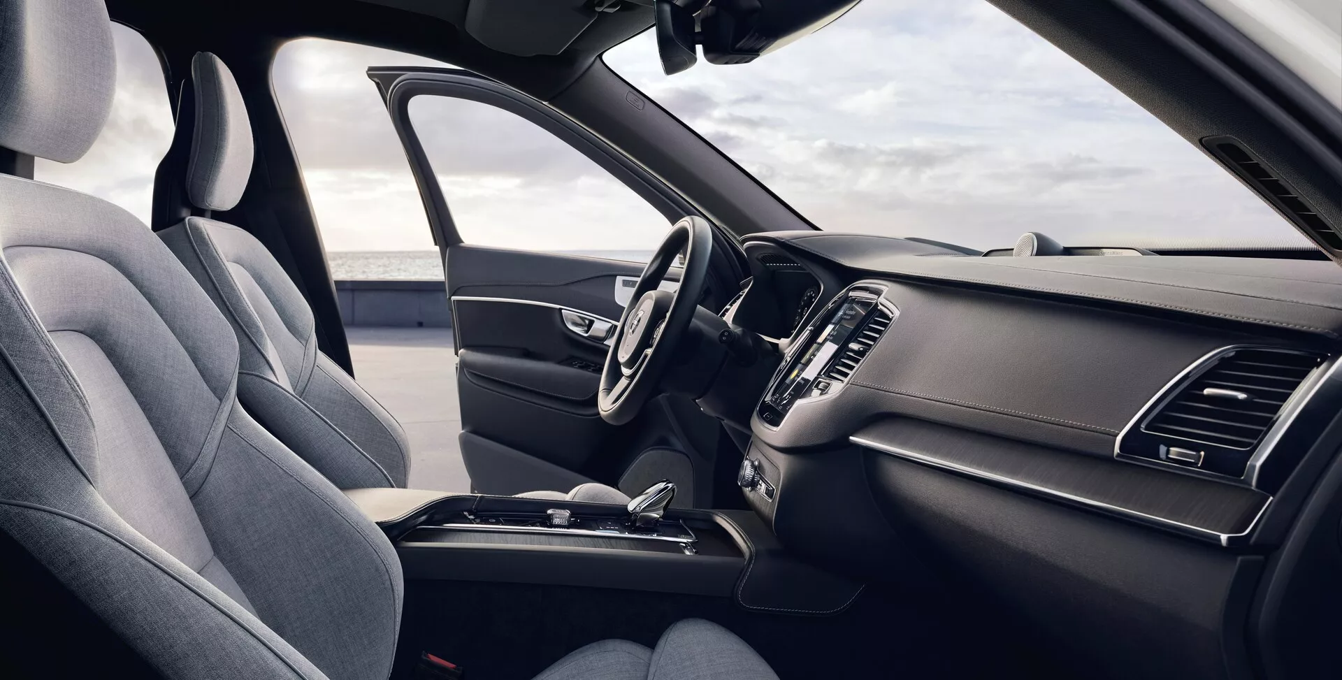 Widok wnętrza Volvo XC90 2015 (2 generacja) z perspektywy przedniego pasażera, przedstawiający ergonomiczne siedzenie kierowcy tapicerowane ciemnym materiałem premium lub skórą syntetyczną, wielofunkcyjna kierownica z zamontowanymi kontrolkami, elegancką deskę rozdzielczą z wyłączonym systemem infotainment z ekranem dotykowym, zintegrowane wloty powietrza, minimalistyczne sterowanie klimatyzacją, centralną konsolę z miejscem do przechowywania, uchwytami na napoje i dźwignią zmiany biegów, wszystko podkreślone jednolitym ciemnym schematem kolorystycznym oświetlonym naturalnym światłem dziennym.