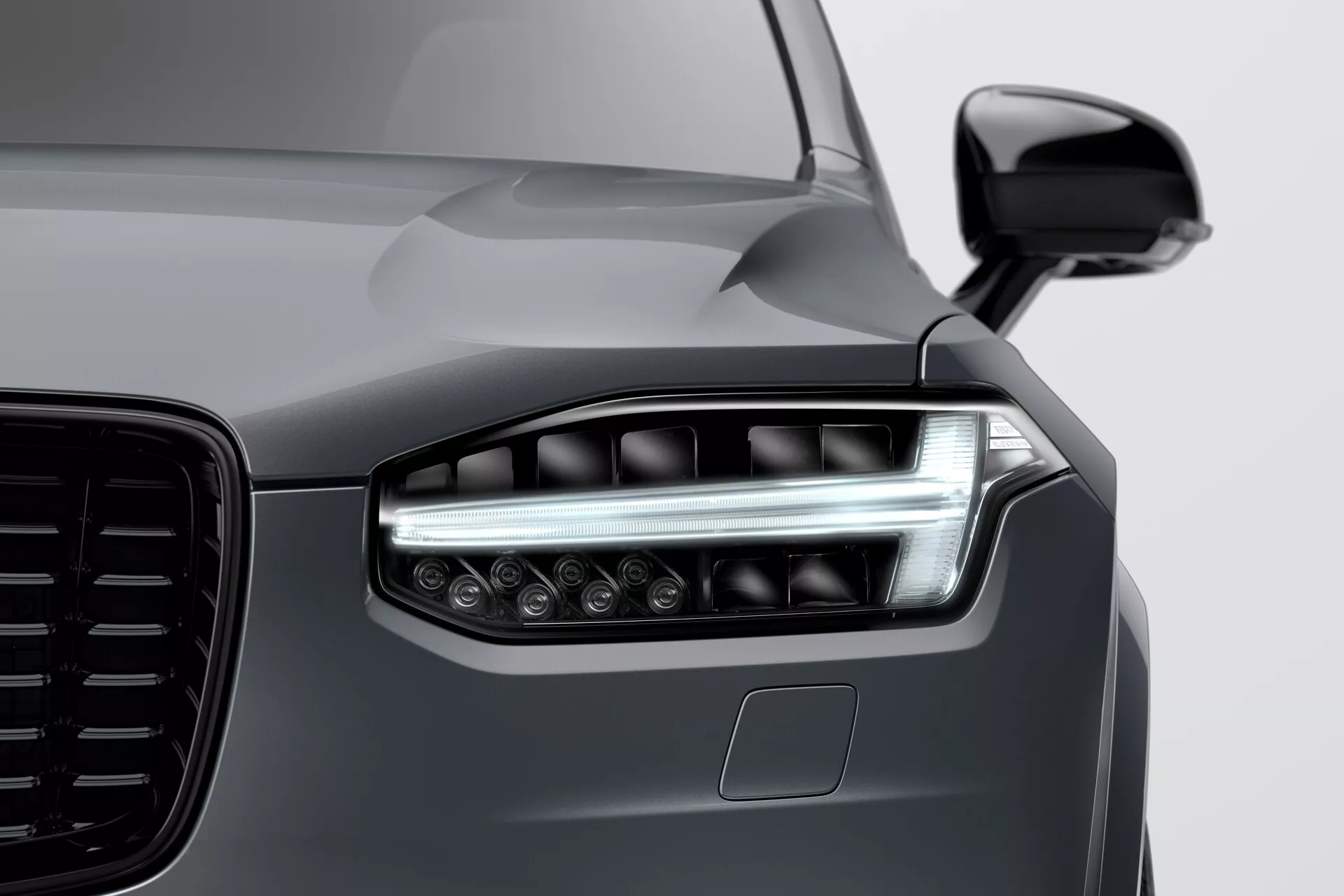 Zbliżenie na przedni róg Volvo XC90 2015 (2. generacja), skupiające się na wyrafinowanym zestawie reflektorów LED, eleganckim pasku światła do jazdy dziennej oraz szczegółowym wzorze grilla. Obraz uchwyca część przedniego zderzaka, potencjalnie z zaawansowanymi czujnikami, oraz aerodynamiczne lusterko boczne z wbudowanymi kierunkowskazami.